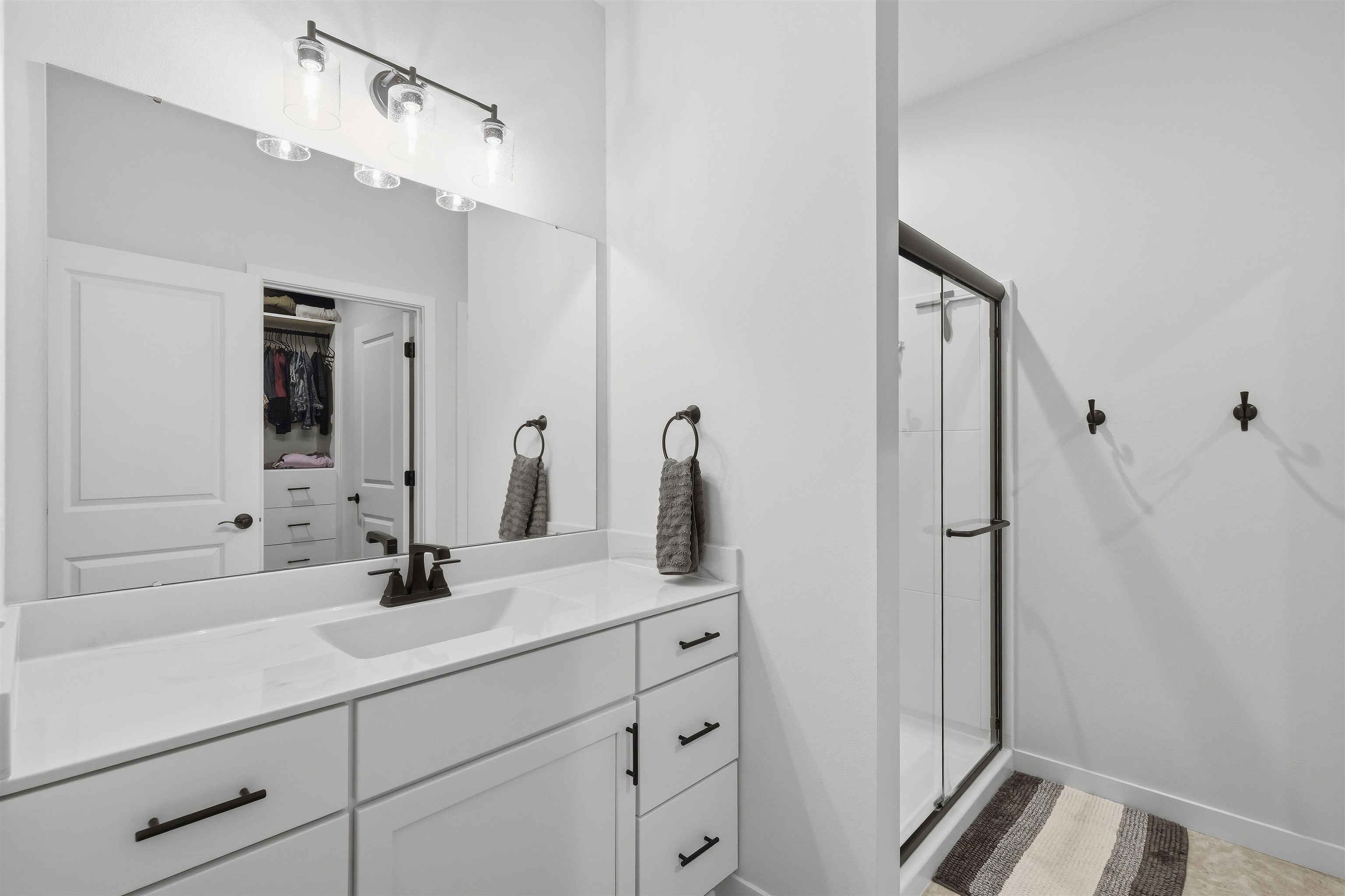 Bathroom with a white vanity and glass shower; an open closet is reflected in the large mirror.