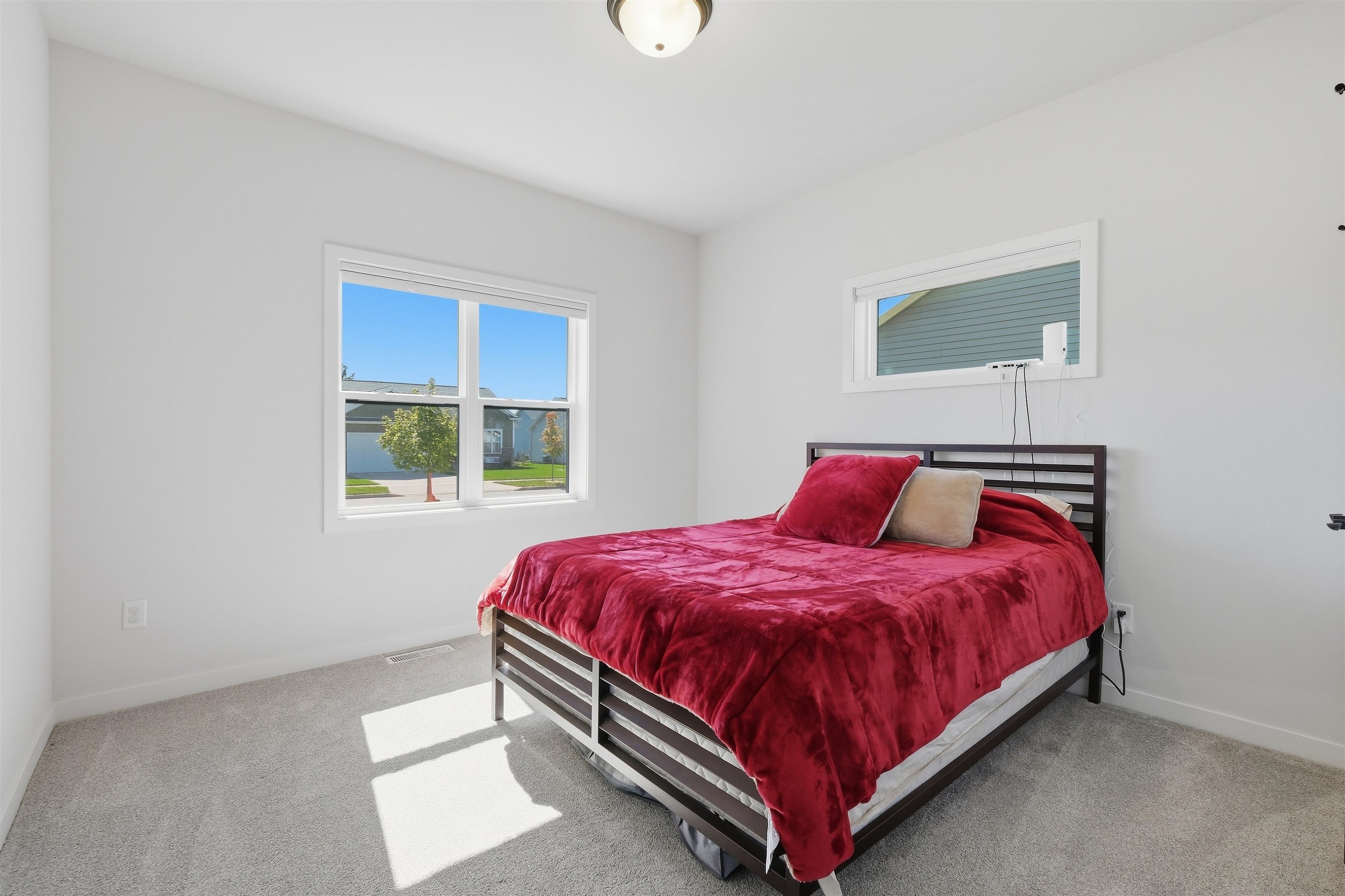 Bedroom with a dark metal bed, deep red bedding, gray carpet, and sunlit windows.
