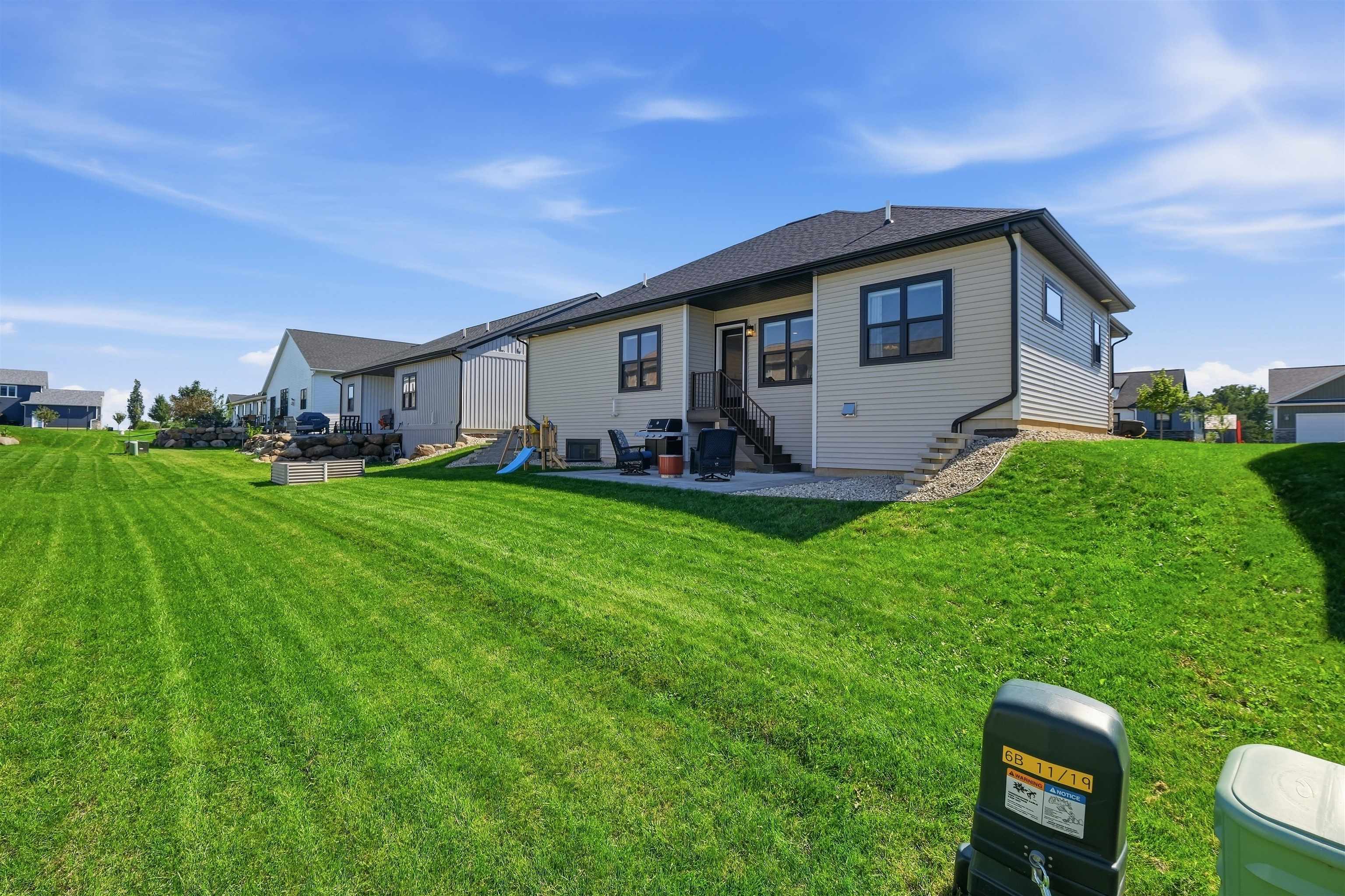 Rear of a gray-sided house overlooking a broad lawn, with deck steps, patio chairs, and a small play structure.