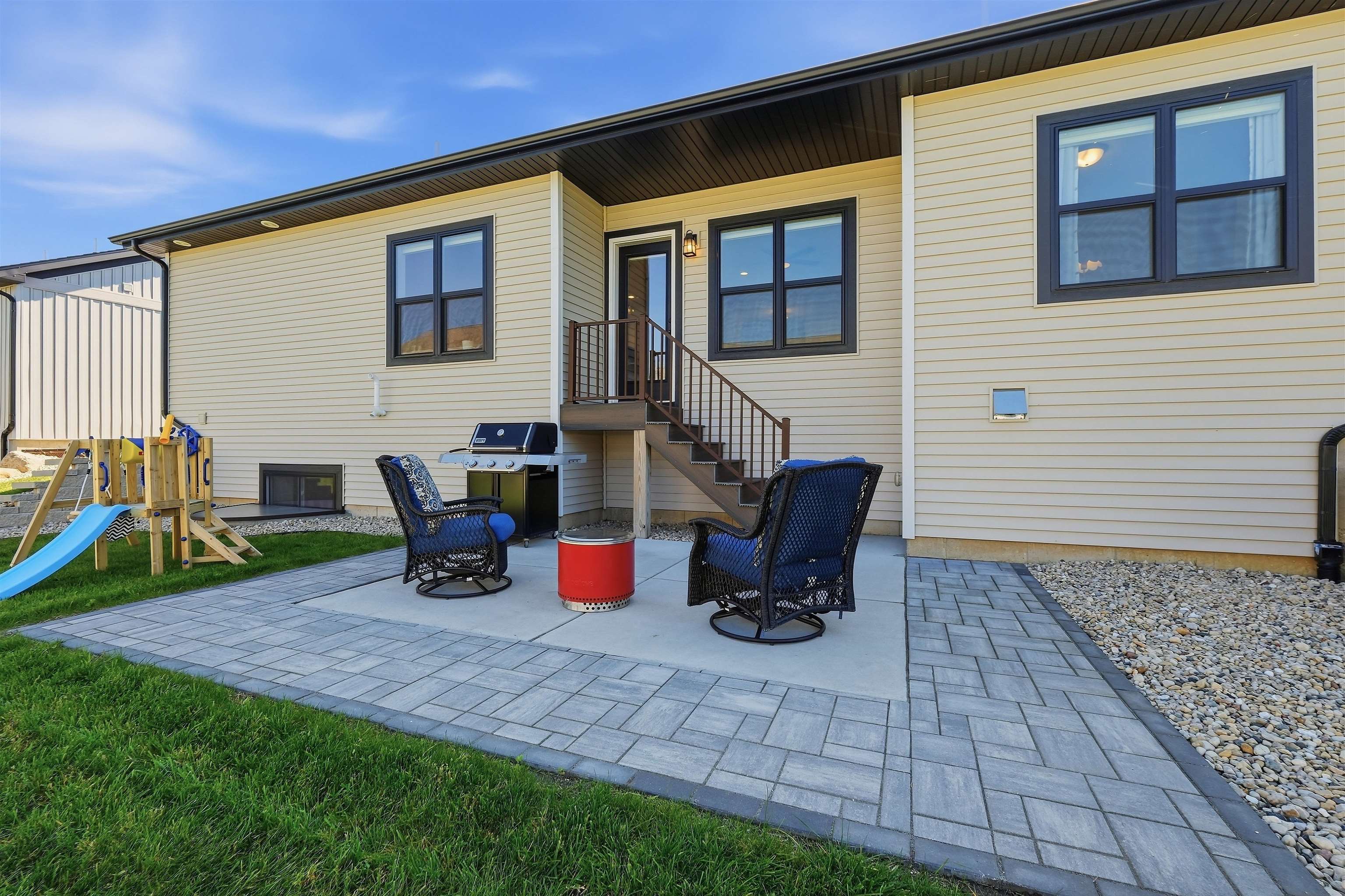 Rear of a beige house with a paved patio, outdoor seating, grill, fire pit, and children’s playset.