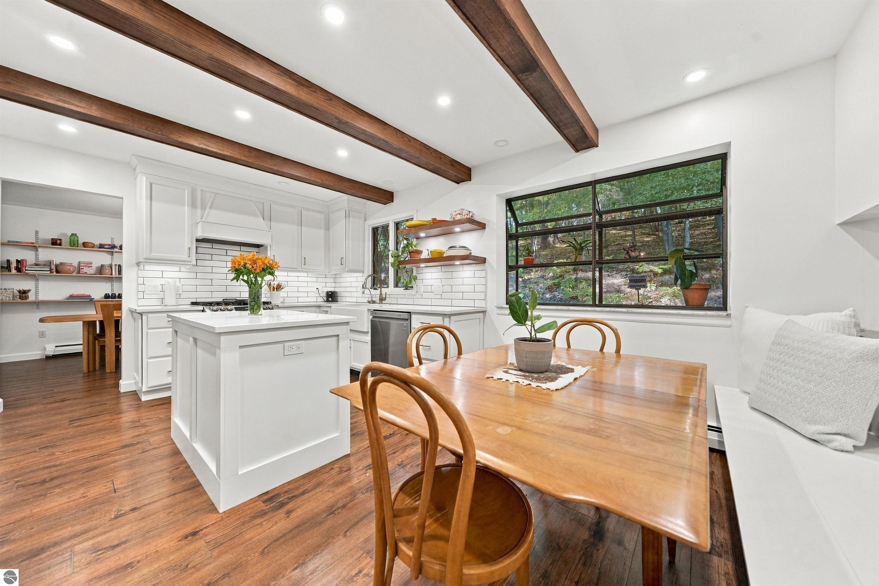 Bright kitchen with a white island, exposed wood beams, and a dining table beside a large window.