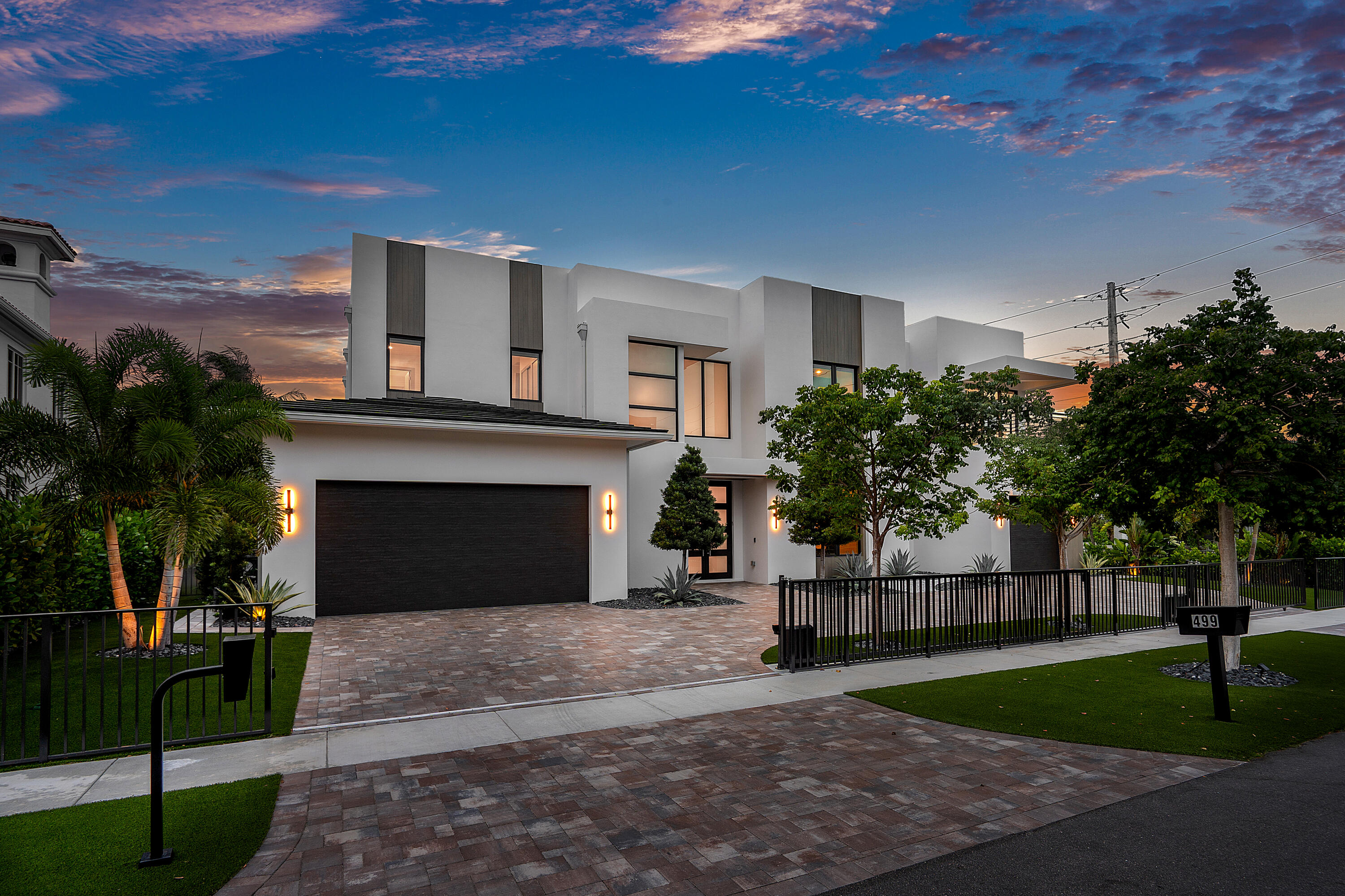 Modern white two-story home with a dark garage, black fence, and palms at dusk.