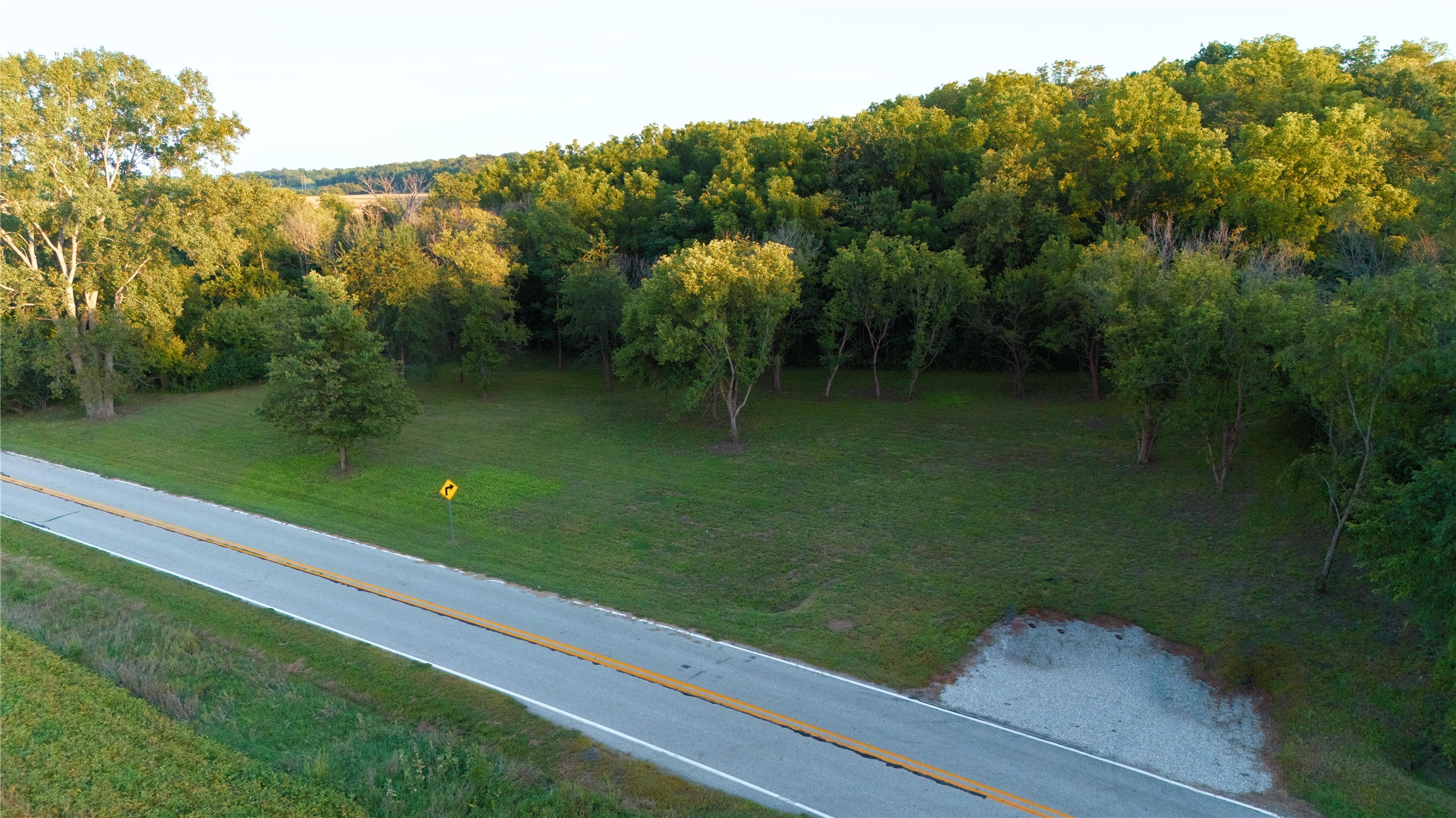 Wooded property beside a two-lane road, with a gravel entrance and grassy clearing.