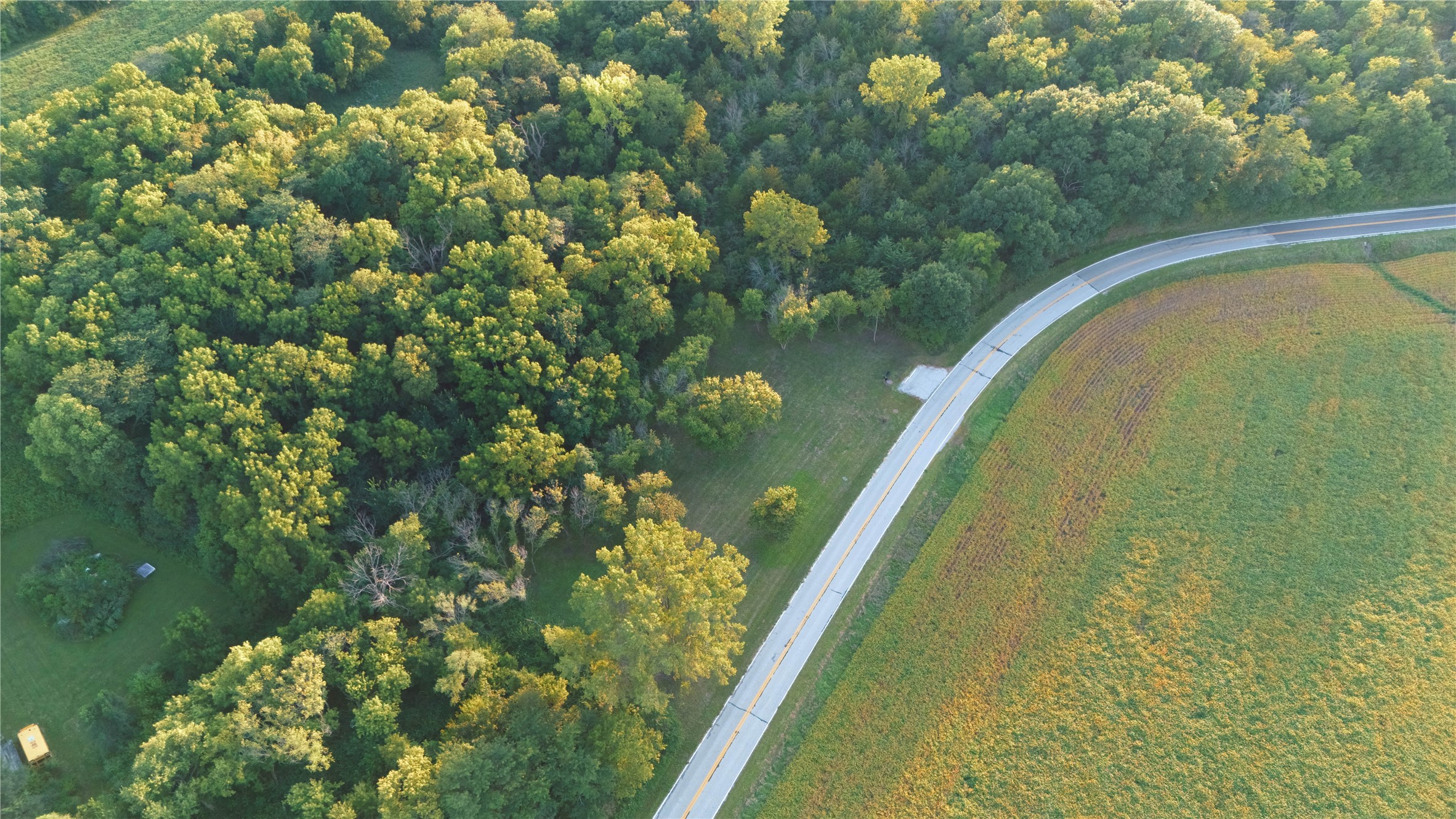 Aerial property view of a curving road bordered by woodland and an open field.