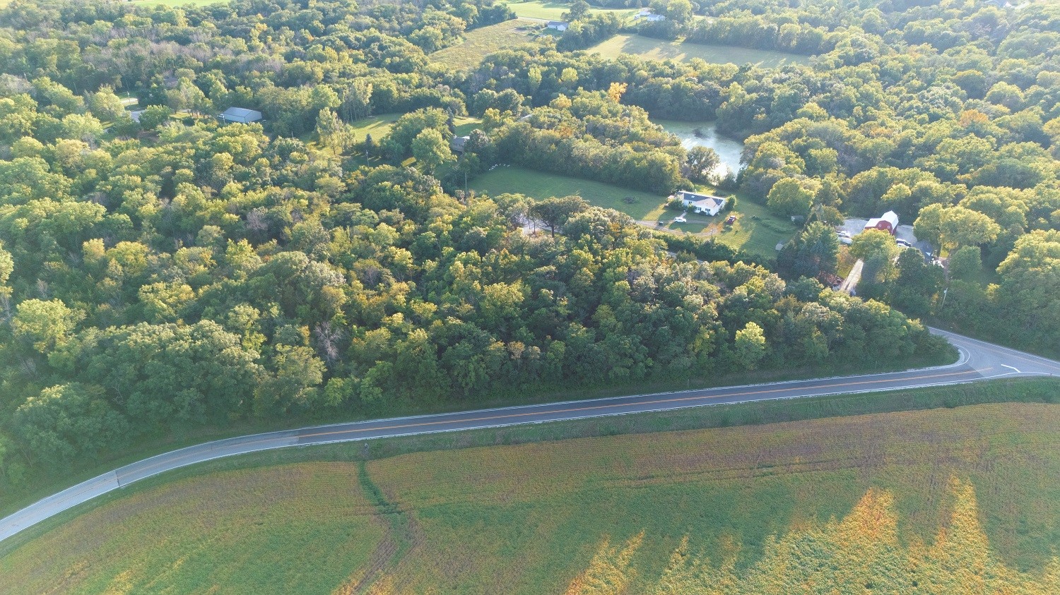Aerial view of wooded acreage with scattered homes, a pond, farmland, and a curving road.