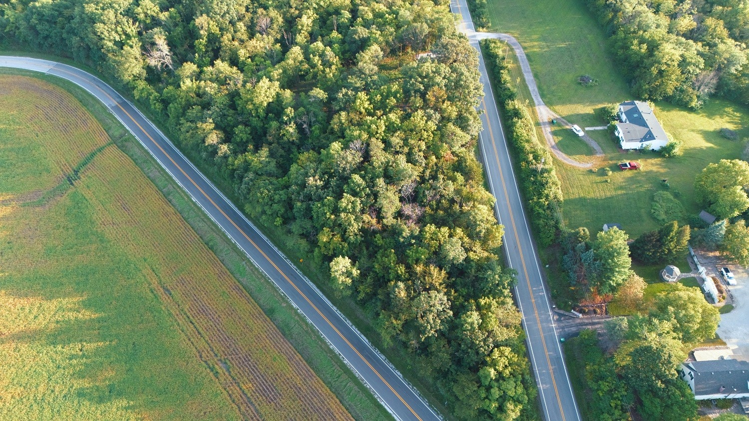 Aerial view of a rural home beside wooded land and fields, with roads along the property.