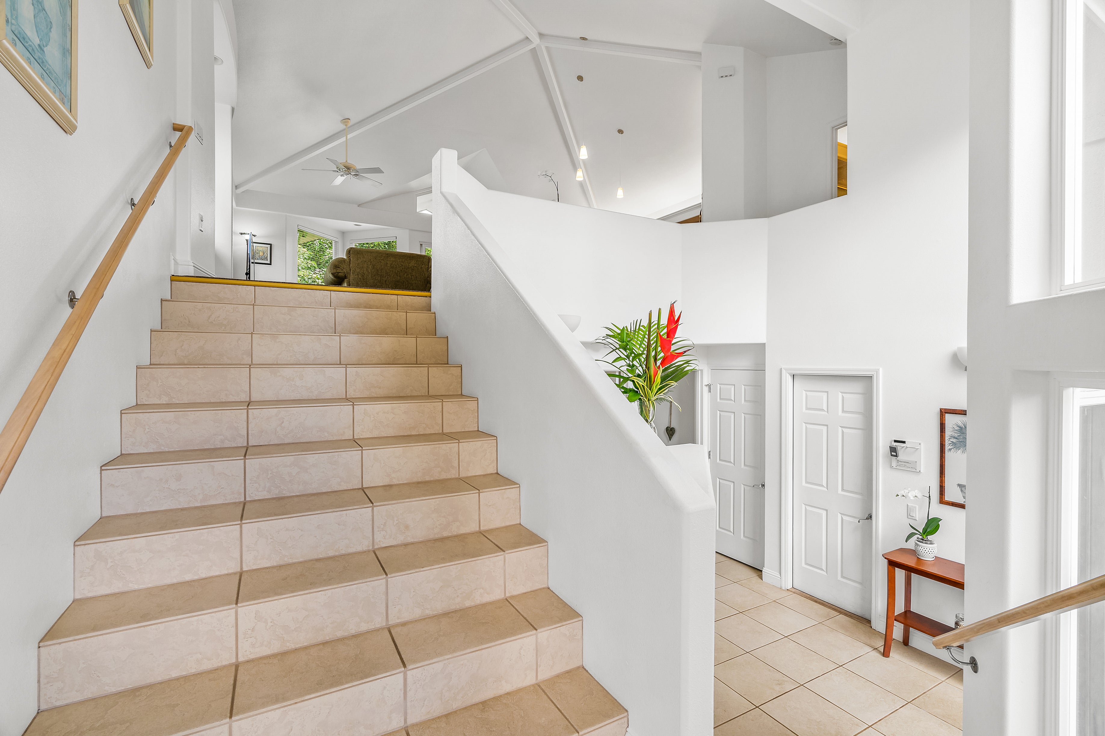 Tiled staircase with a wood handrail rises beside a bright, open foyer and tropical plant.