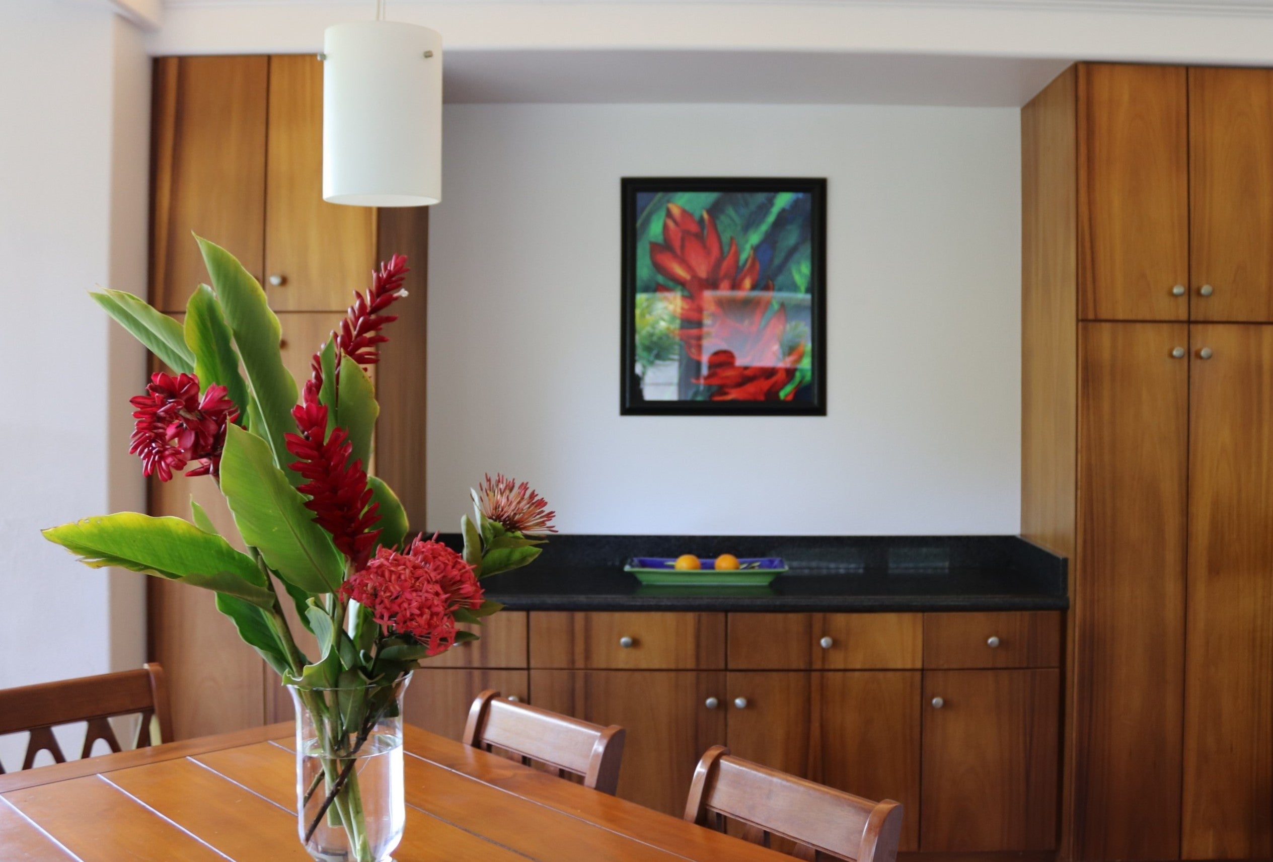 Dining area with warm wood cabinetry, a table, and a vase of red tropical flowers.