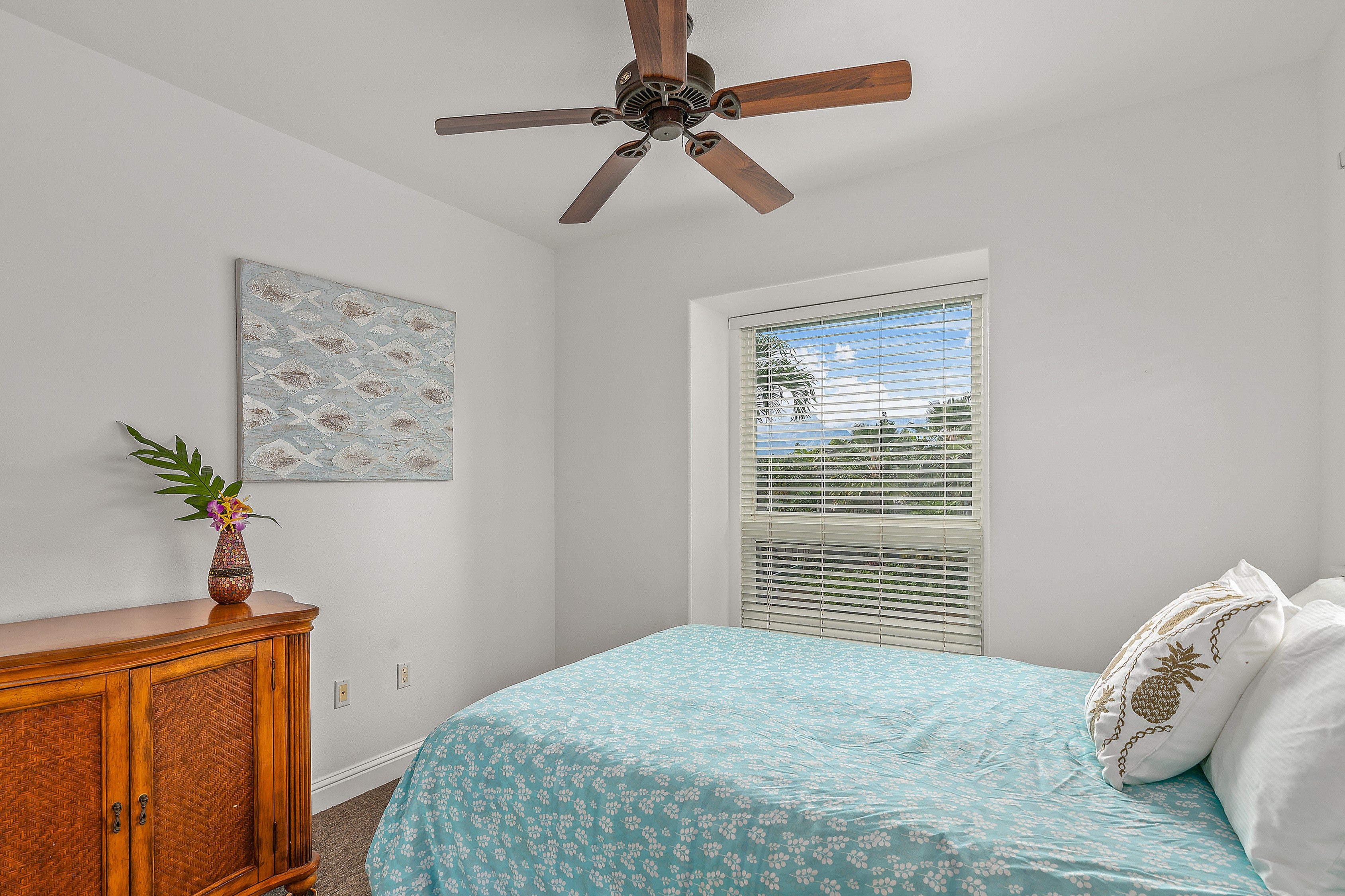 Bedroom with a turquoise floral bedspread, wooden cabinet, and window overlooking tropical greenery.