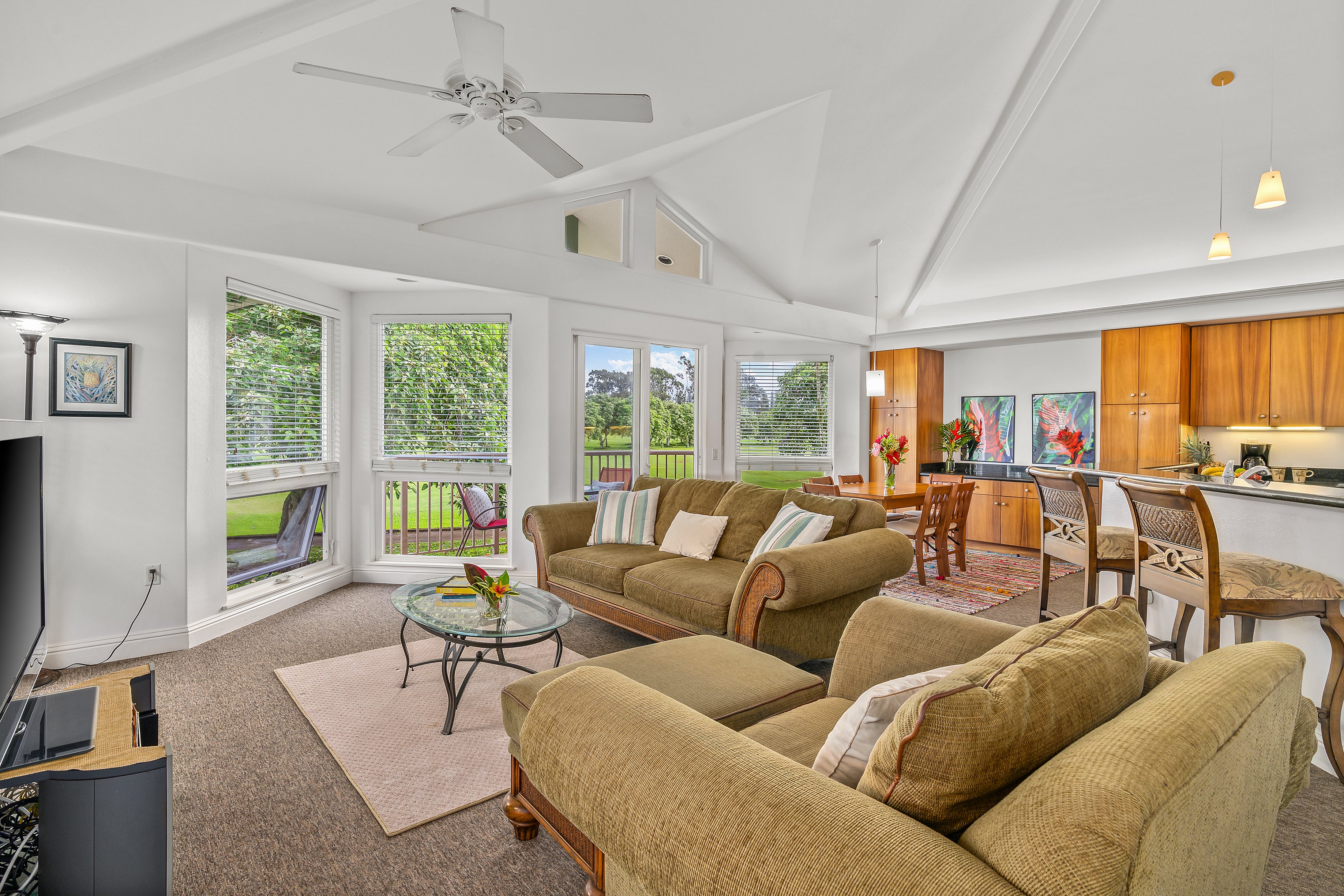 Furnished living room with vaulted ceiling, tan sofas, wood kitchen cabinets, and windows overlooking a green lawn.