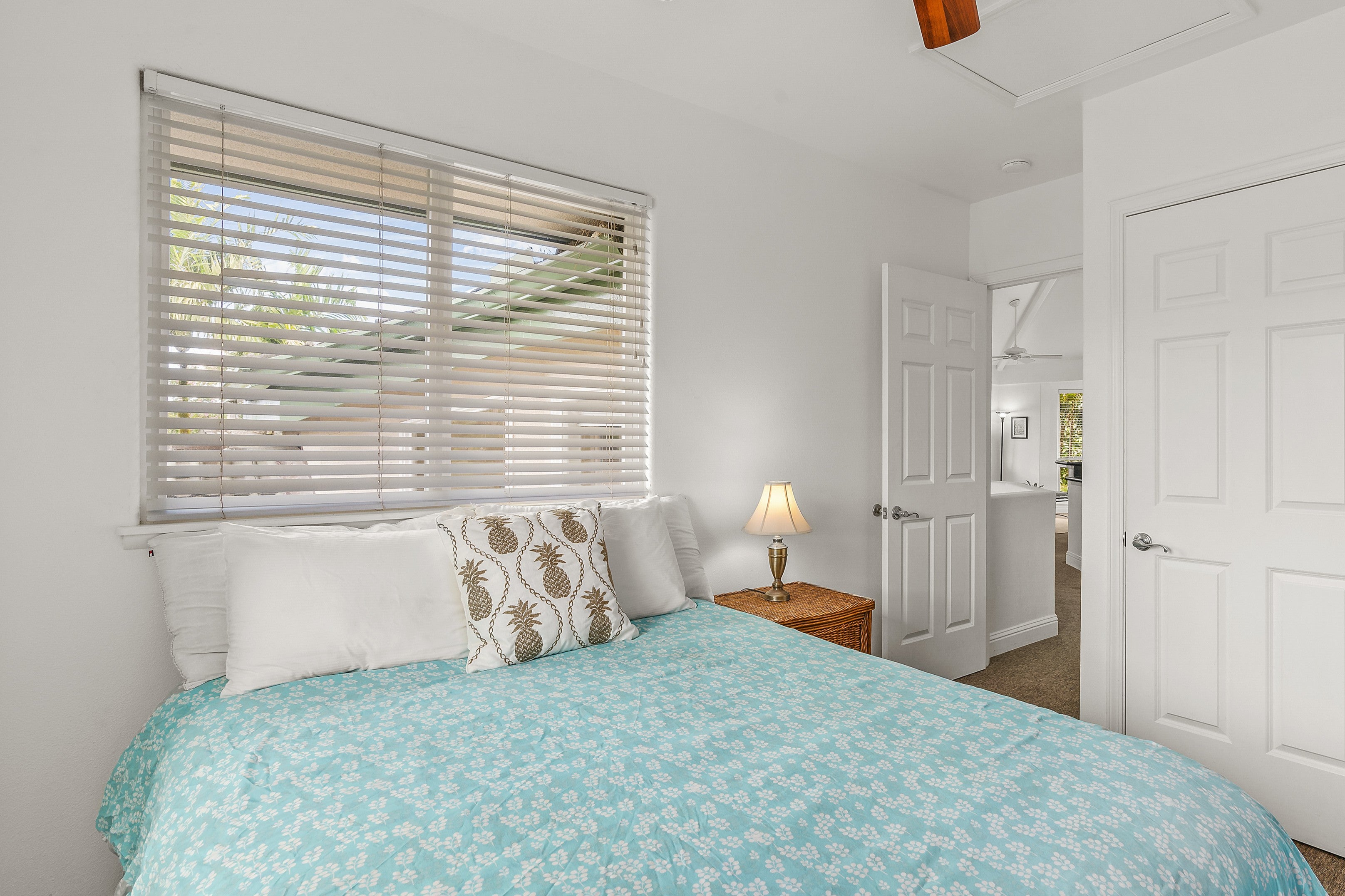 Bedroom with a turquoise floral bedspread, pineapple-print pillow, and window overlooking tropical greenery.