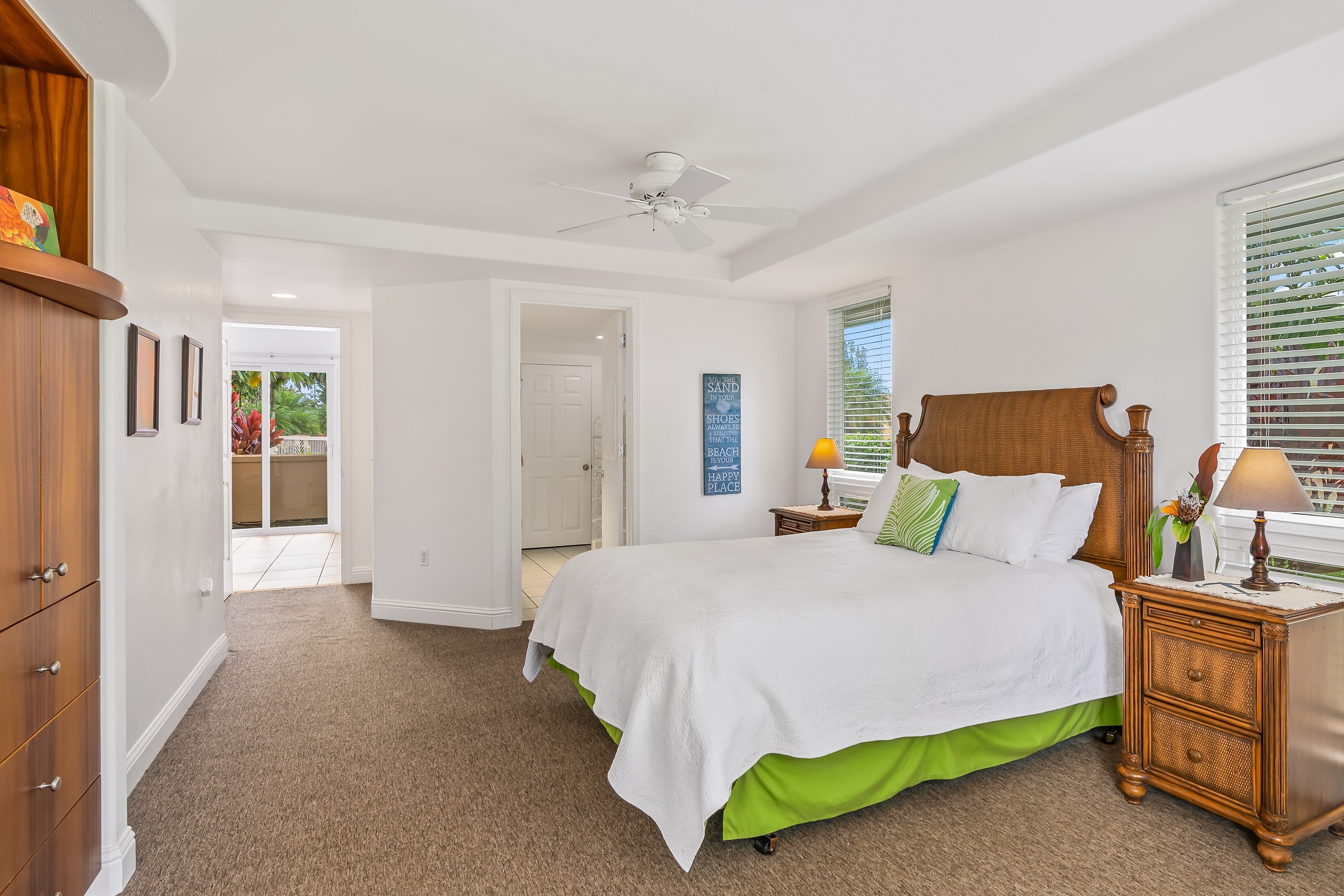 Furnished bedroom with a white-covered bed, bright green bed skirt, carved wooden headboard, and tropical foliage outside.