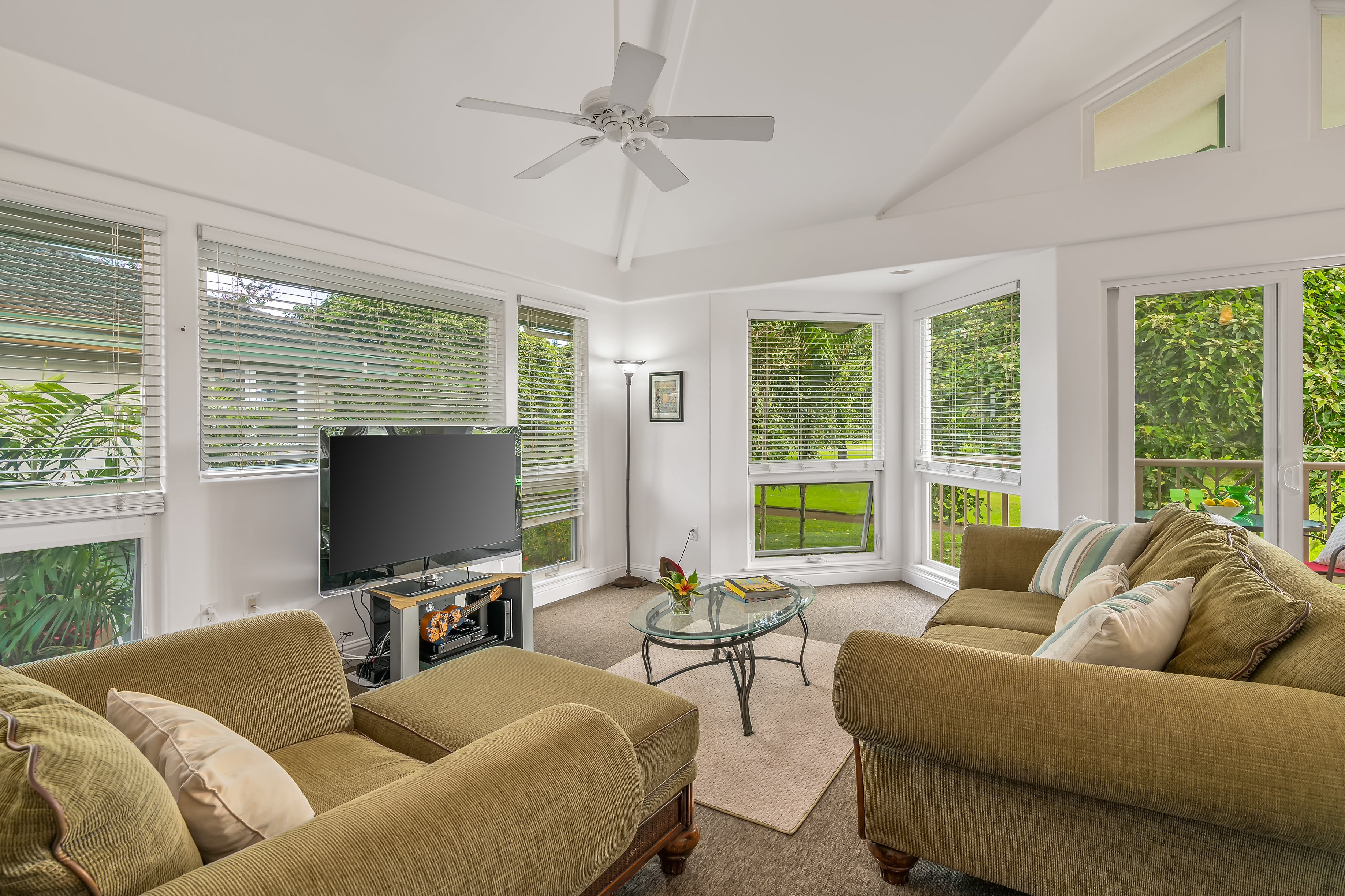 Furnished living room with olive sofas, a glass coffee table, and large windows overlooking greenery.