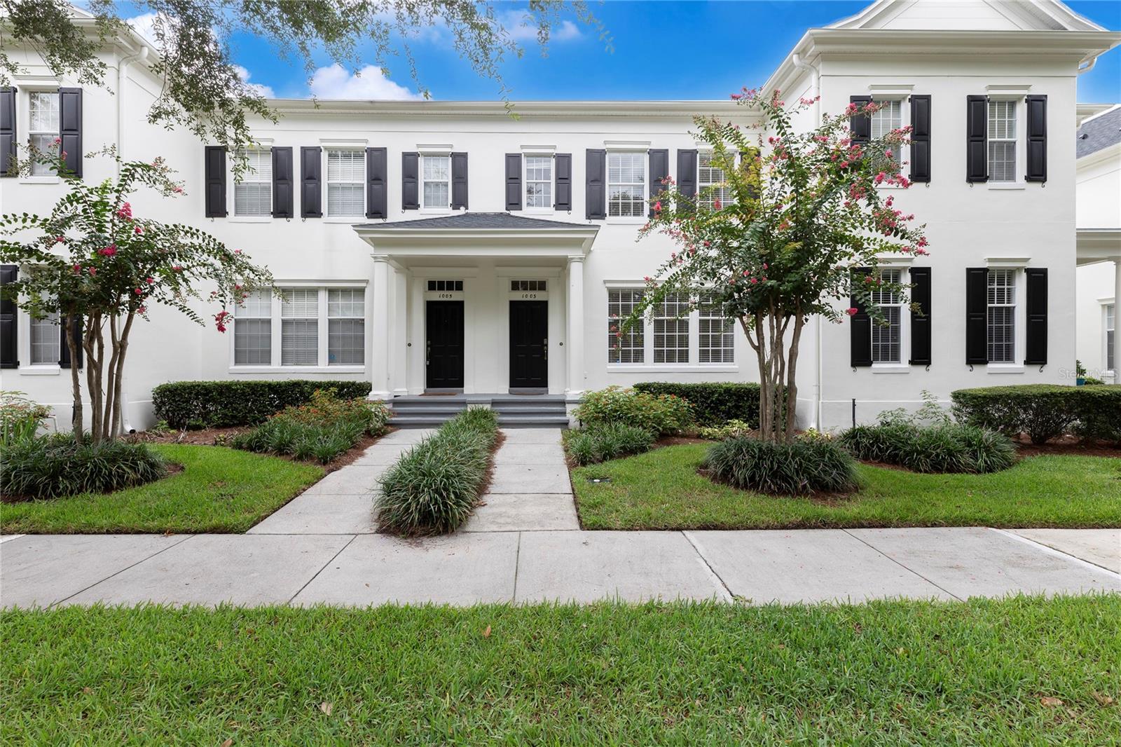 White two-story residence with black shutters, paired entry doors, flowering trees, and a front walkway.