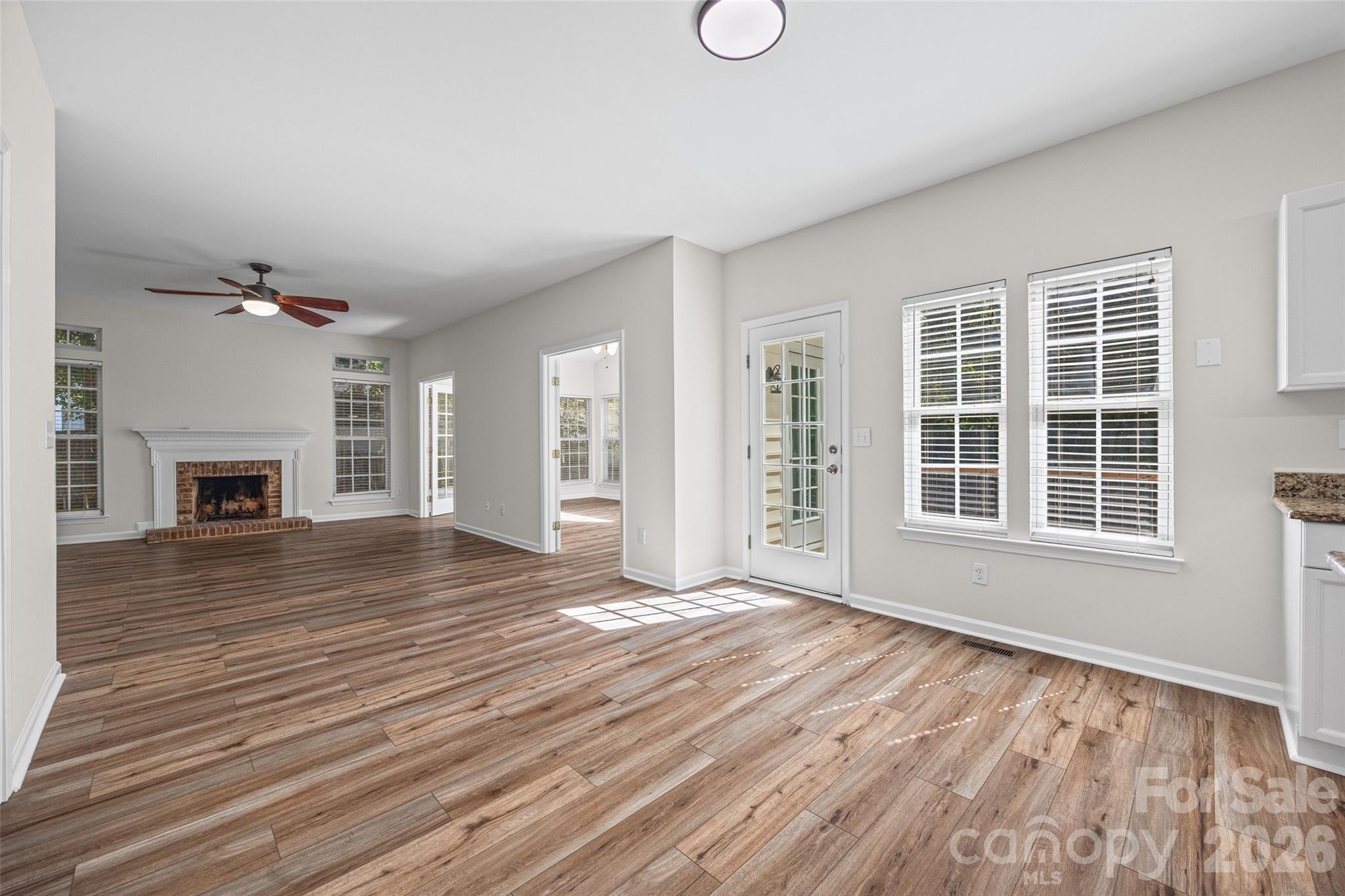 Vacant living room with wood-look flooring, brick fireplace, ceiling fan, and glass-paneled door.