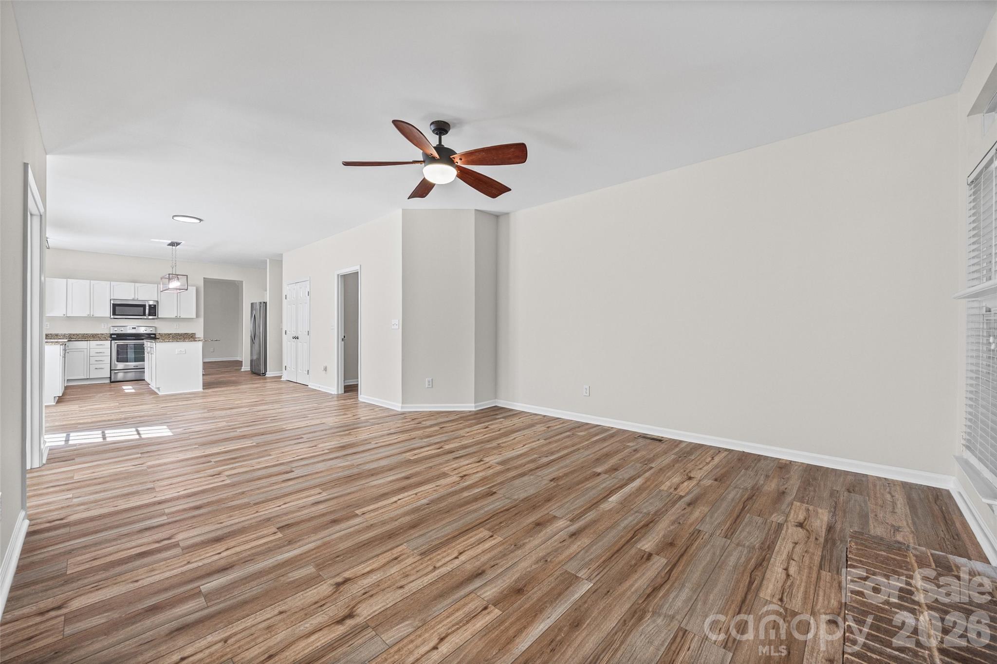 Open living area with wood-look flooring, ceiling fan, and white kitchen with stainless steel appliances.
