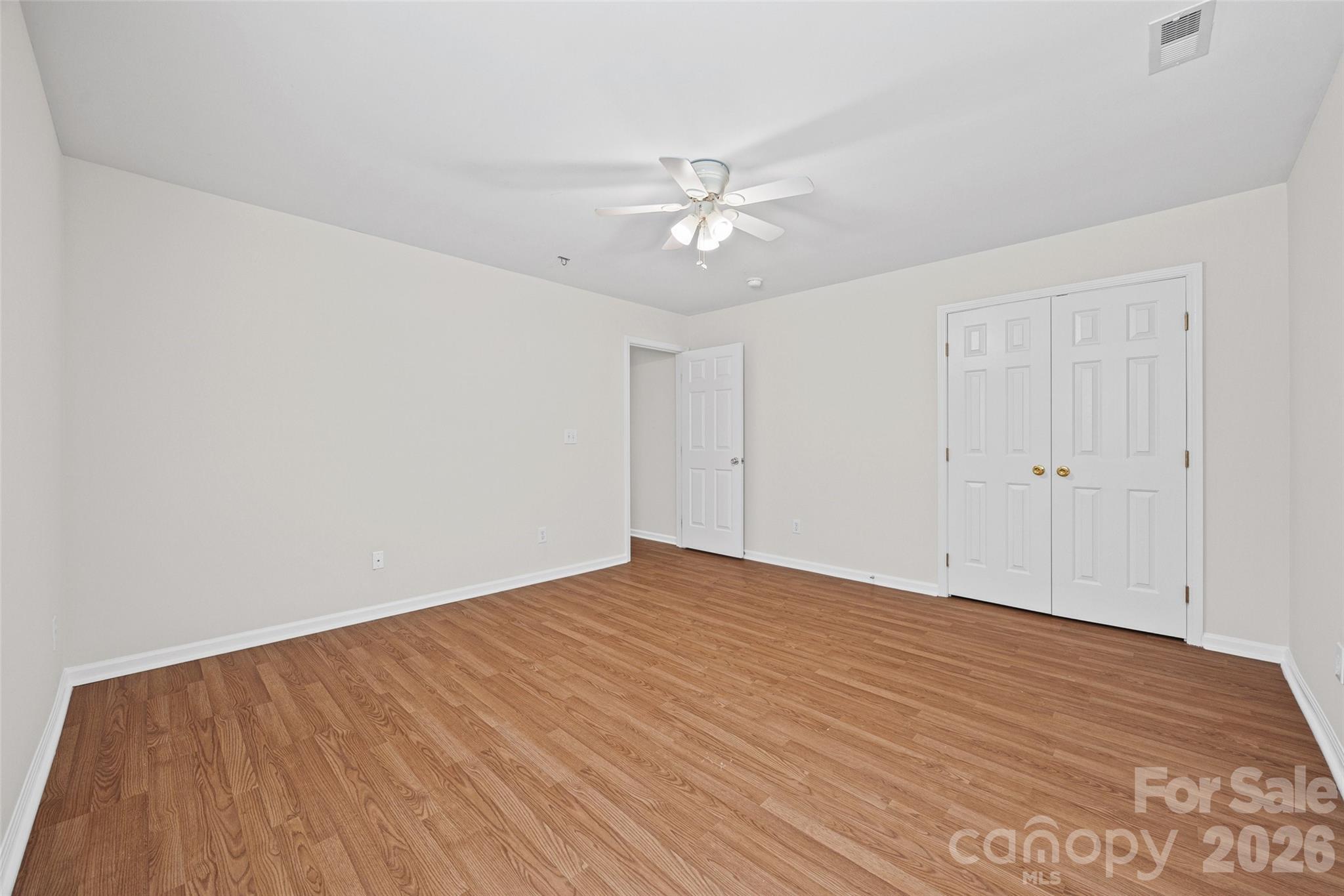 Vacant bedroom with warm wood flooring, a white ceiling fan, and double closet doors.