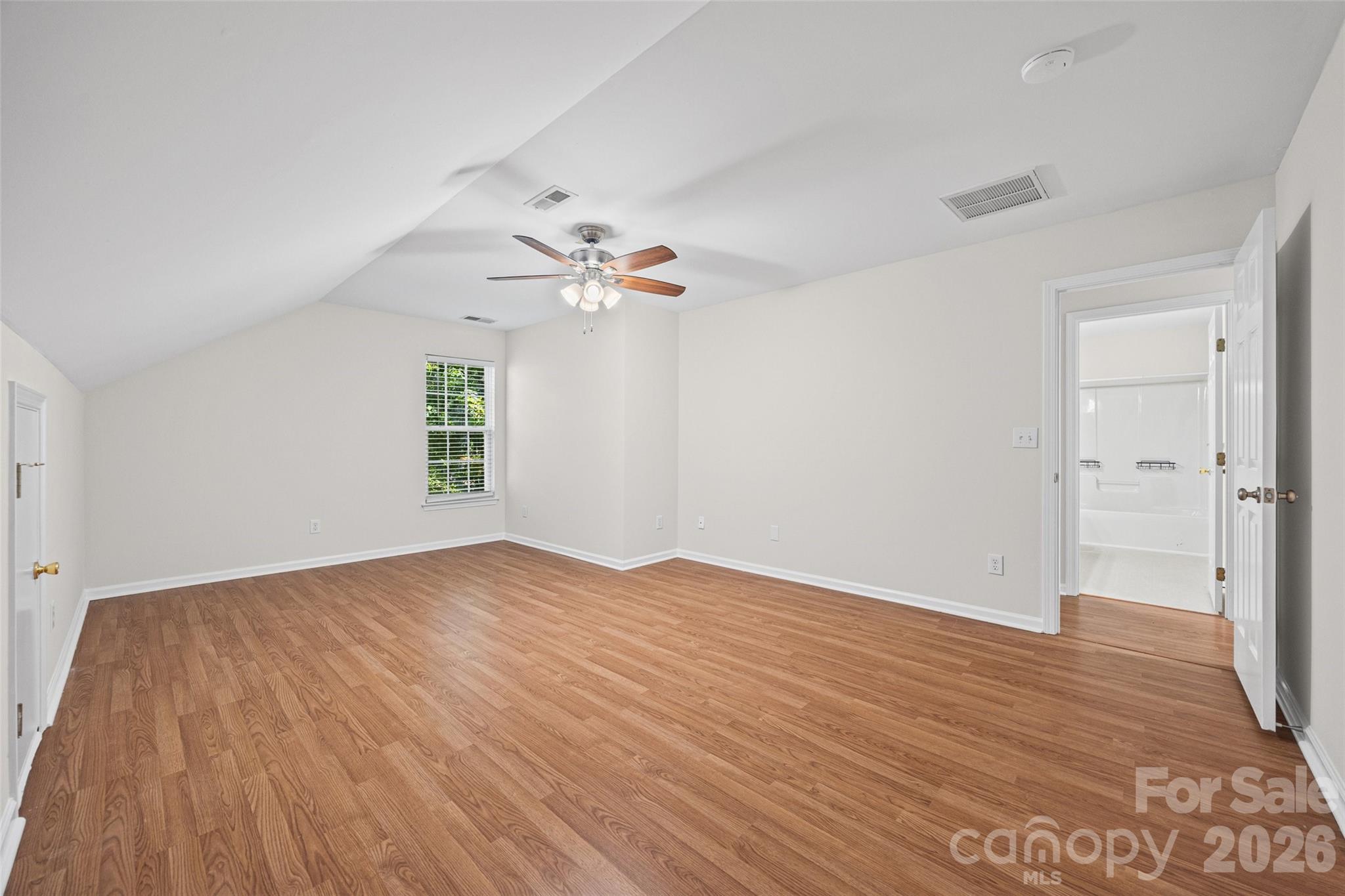 Empty bedroom with wood floors, vaulted ceiling, ceiling fan, and an open doorway to the bathroom.