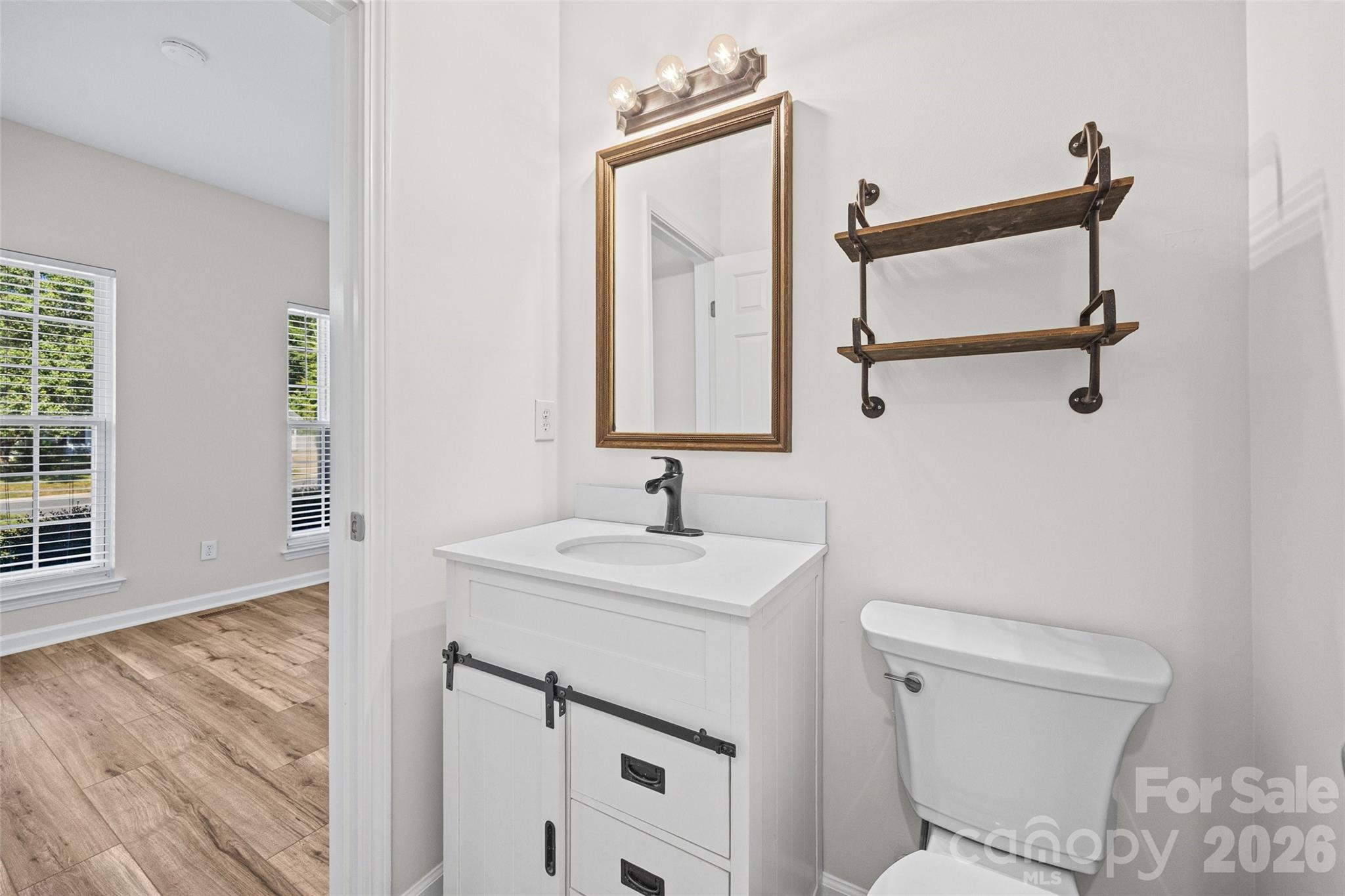 Bathroom with white vanity, framed mirror, wooden shelves, and toilet beside an open doorway.