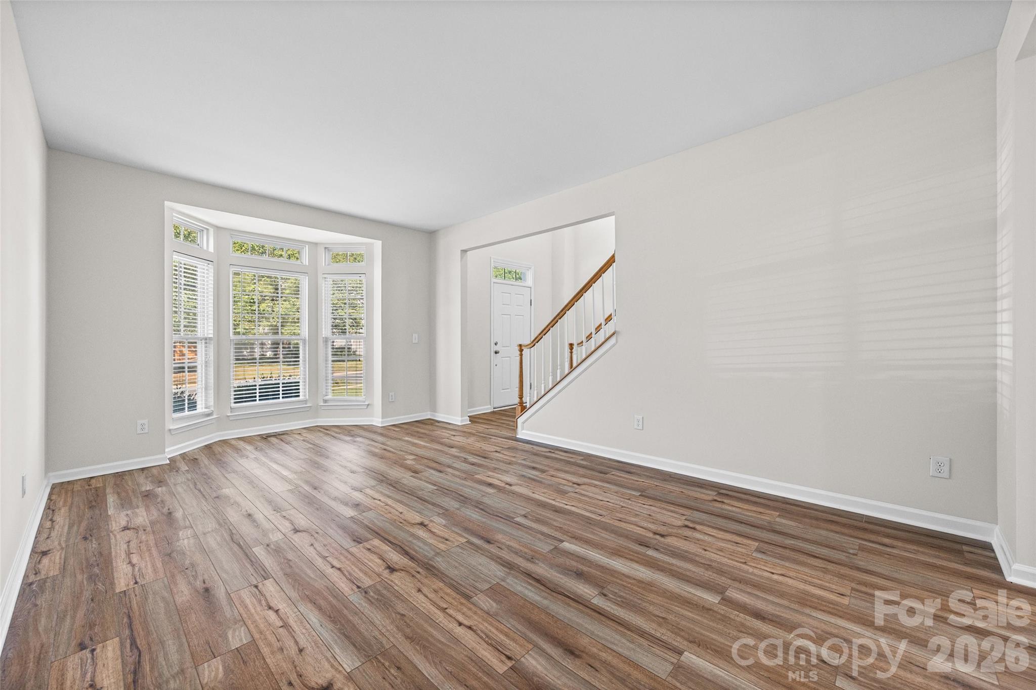 Empty living room with wood-look floors, bay windows, and a staircase beside the front entry.