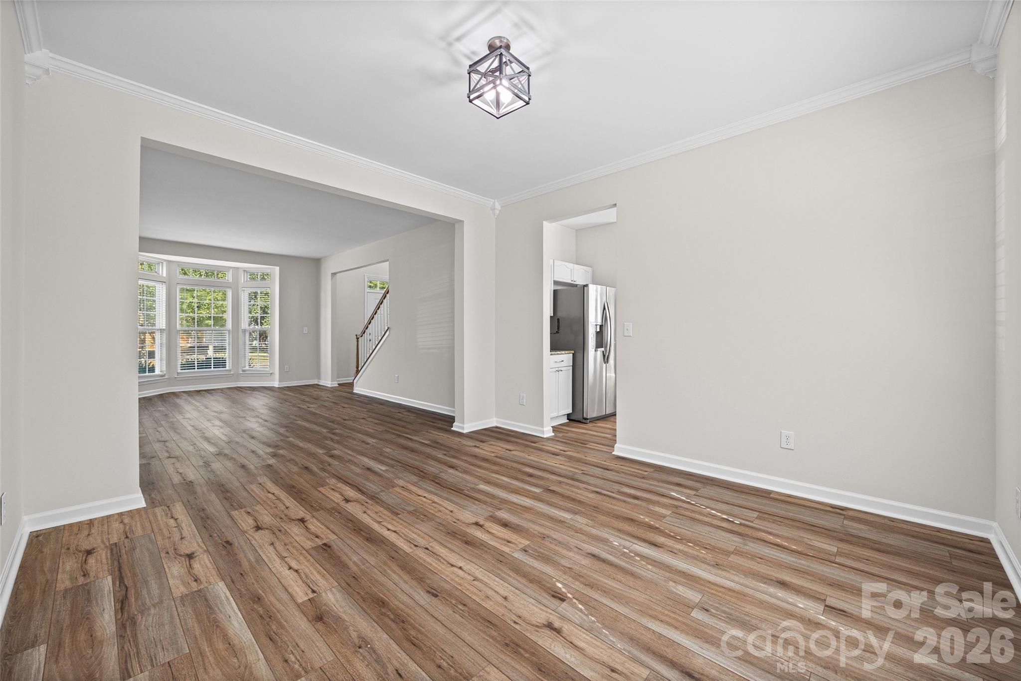Vacant open-plan living area with wood-look floors, tall windows, staircase, and stainless steel refrigerator.