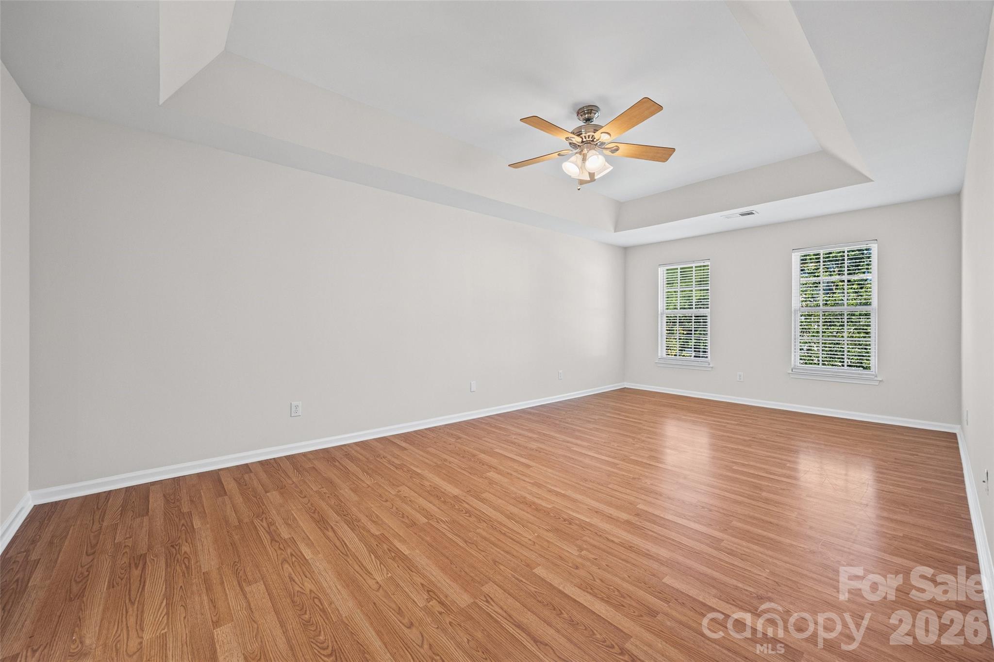 Vacant bedroom with hardwood floors, a tray ceiling, and two windows with white blinds.