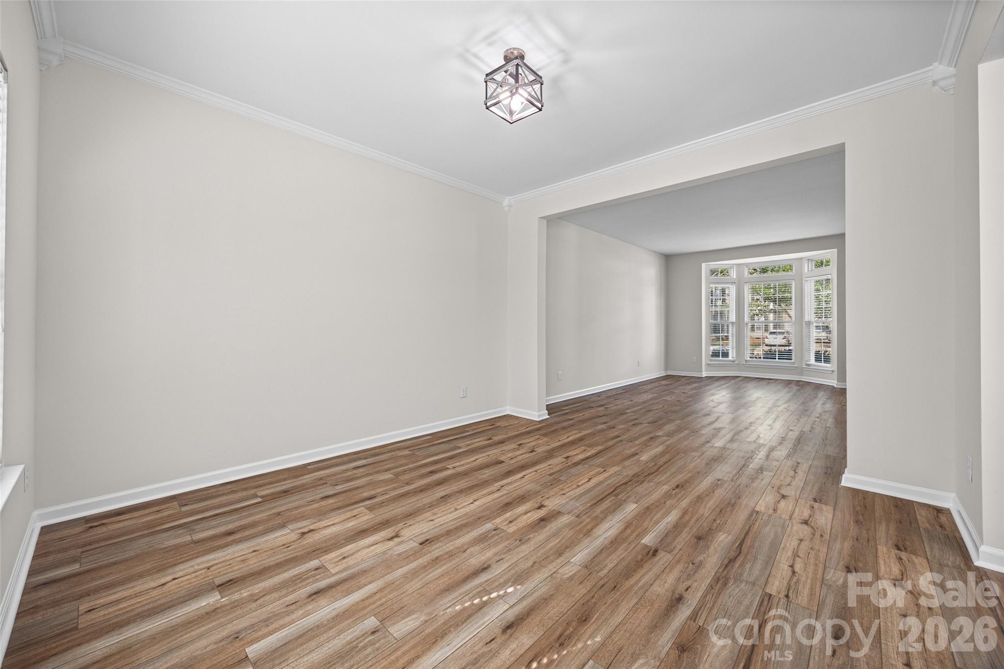 Empty room with wood-look flooring and a wide opening to a bright bay-window room.