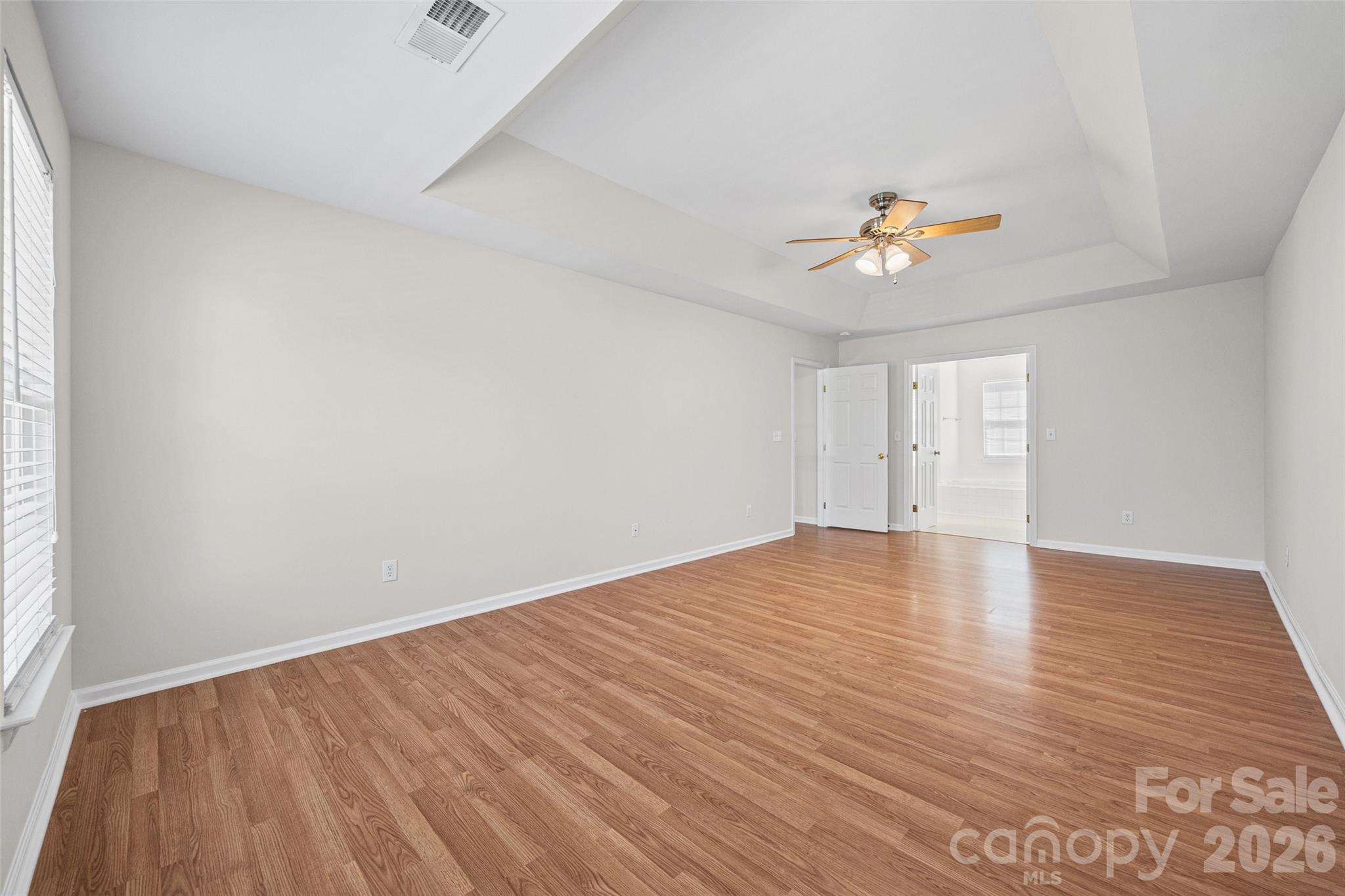 Empty bedroom with hardwood floors, a tray ceiling, ceiling fan, and an open doorway to the bathroom.