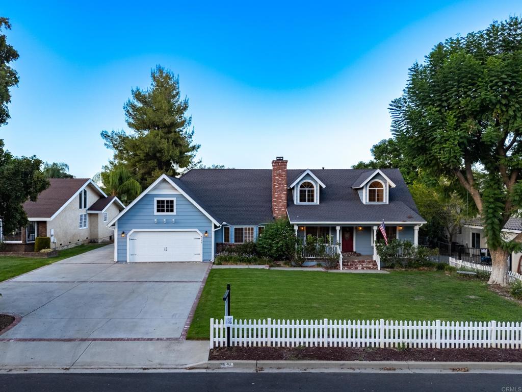 Blue house with dormer windows, brick chimney, wide driveway, front lawn, and white picket fence.