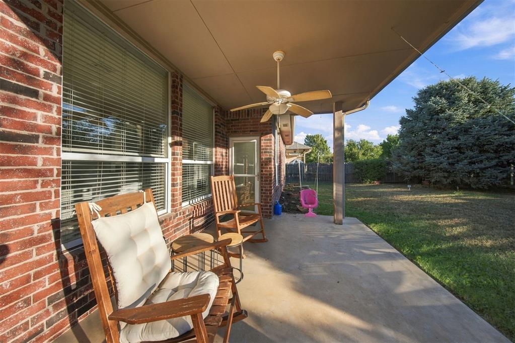 Covered brick patio with a ceiling fan and wooden chairs overlooking a grassy backyard.