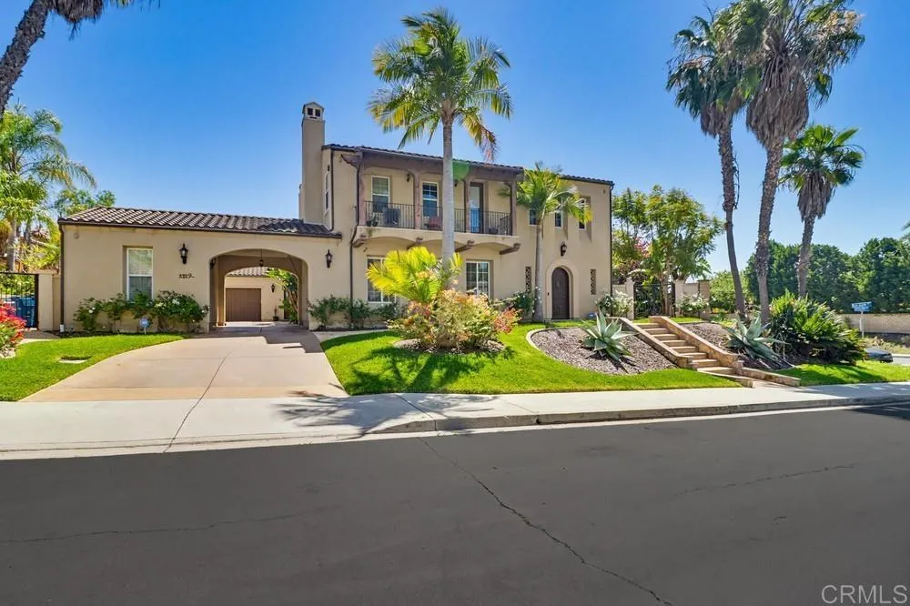 Two-story stucco home with an arched driveway passage, balcony, and palm-lined landscaped yard.