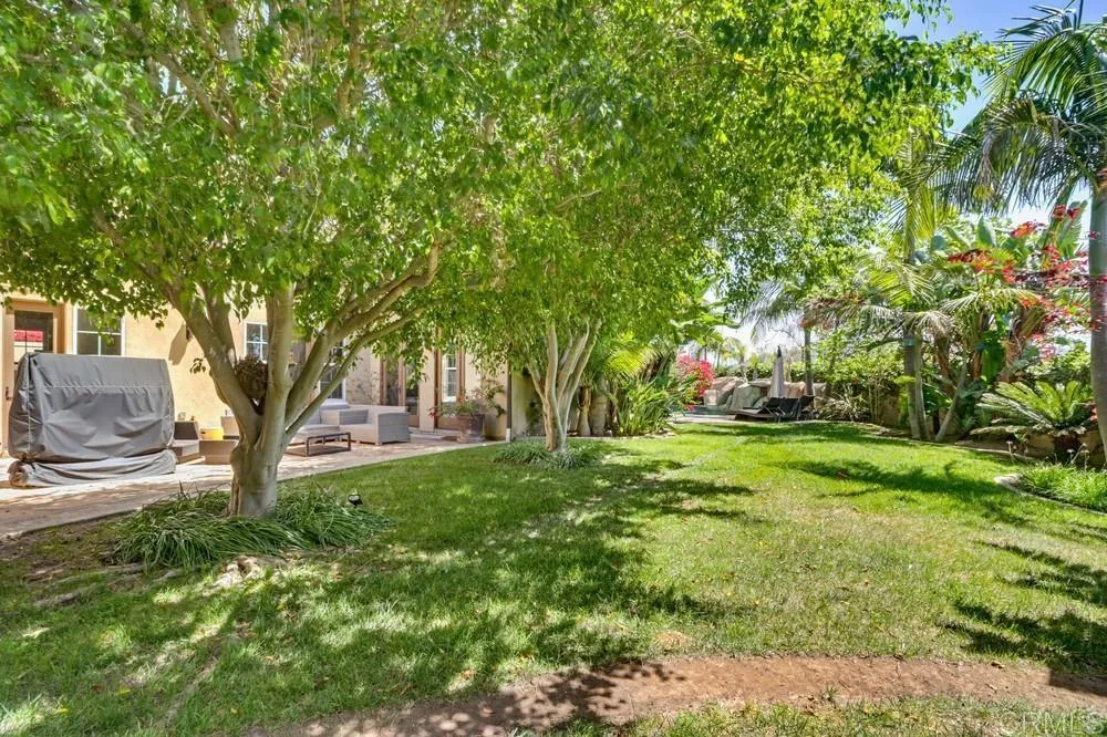 Backyard lawn shaded by mature trees, with tropical landscaping and a furnished patio.