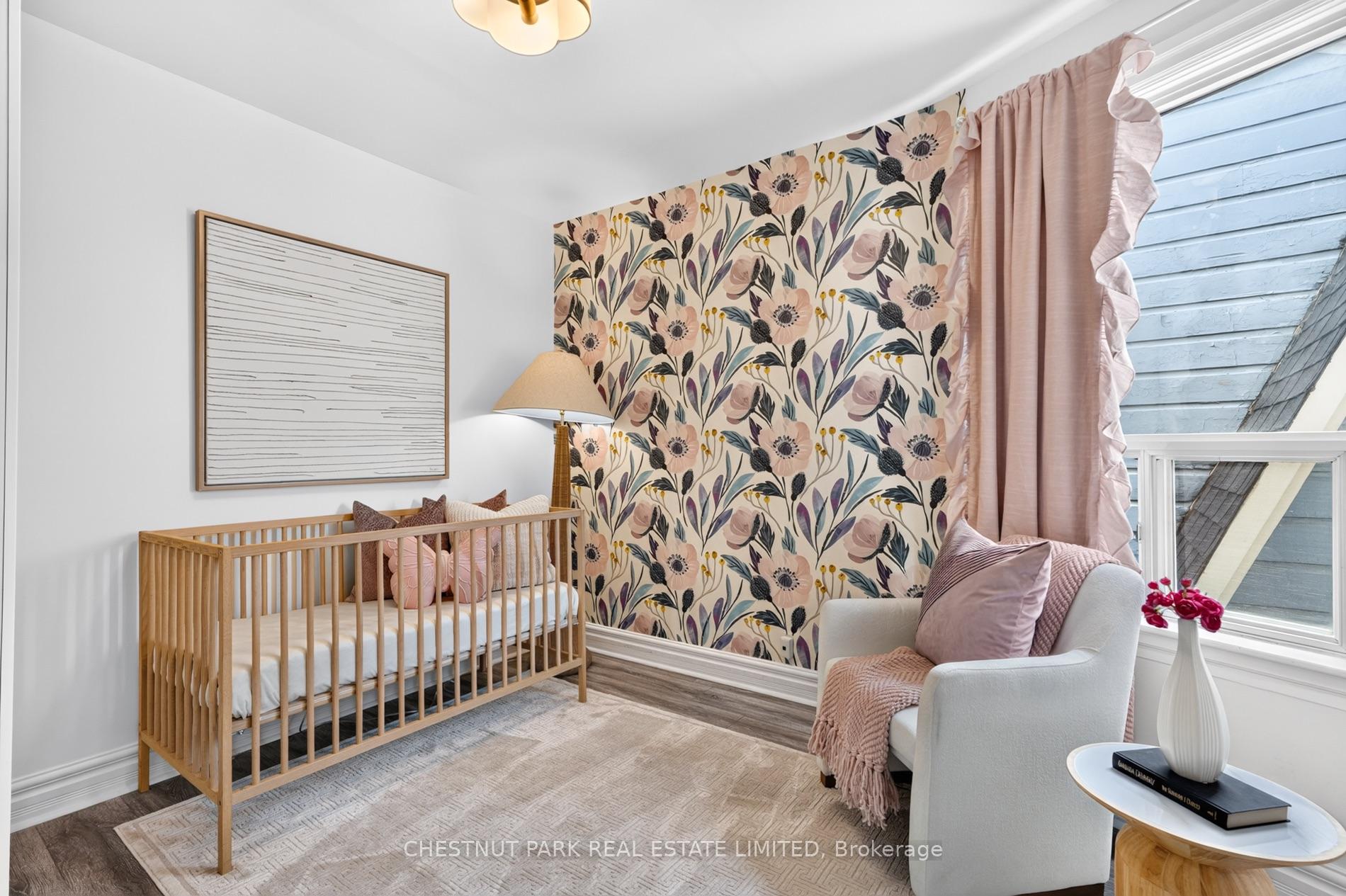Nursery with a wooden crib, floral wallpaper, and a white armchair beside a large window.
