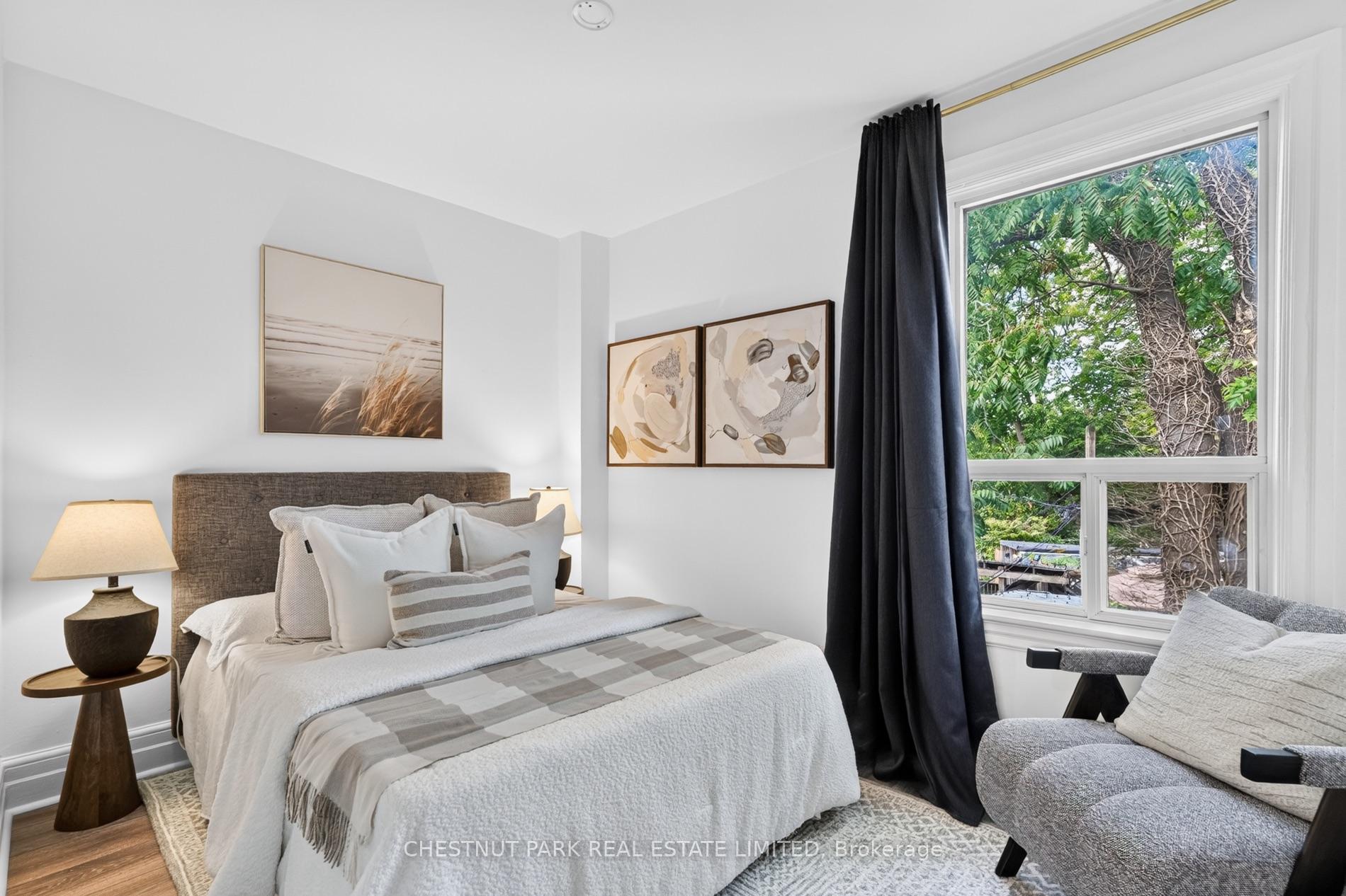 Bright staged bedroom with white and gray bedding, black curtains, and a large window overlooking trees.