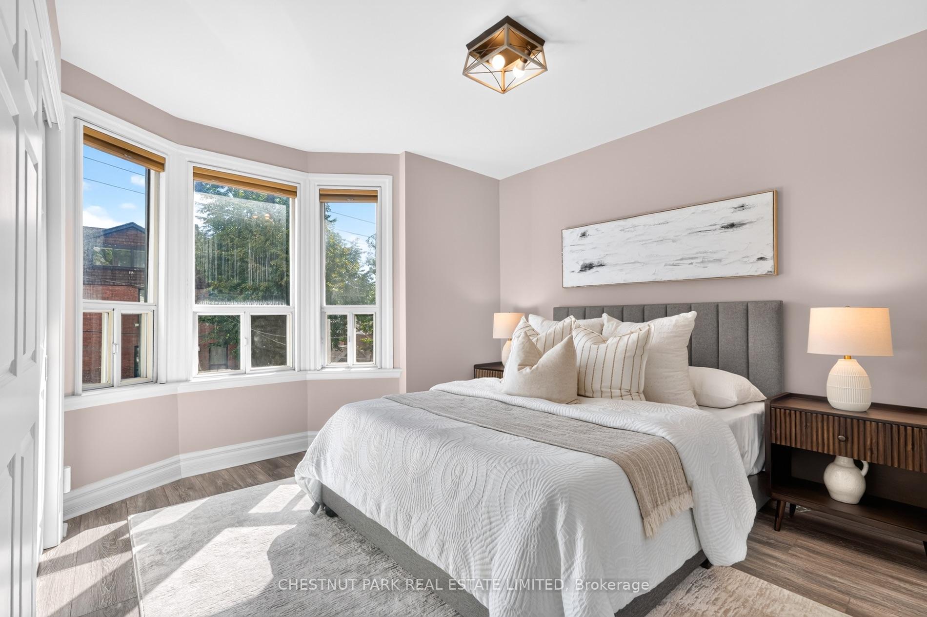 Staged bedroom with white bedding, gray headboard, and three tall bay windows.