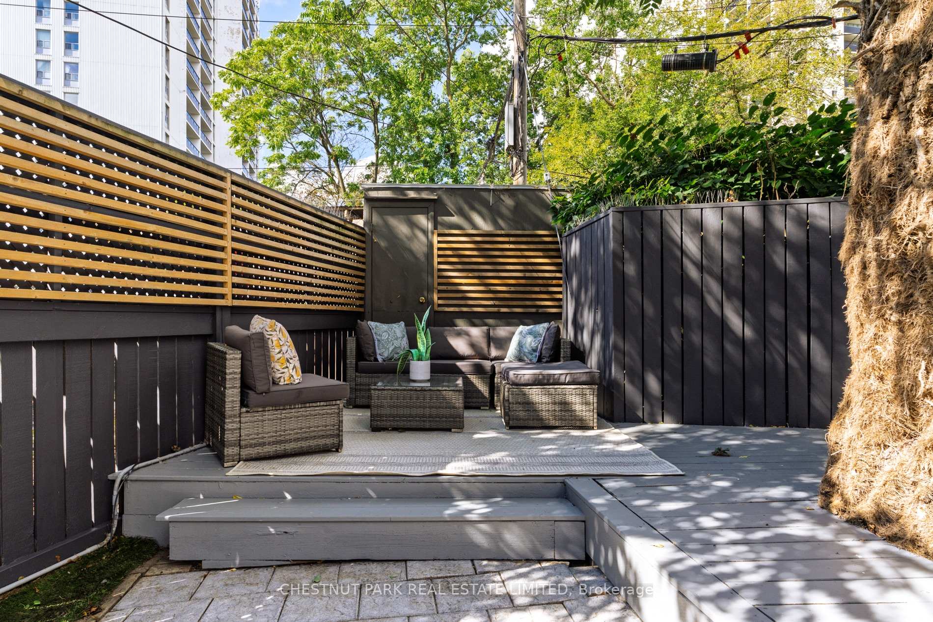 Backyard patio with woven seating, gray decking, wood privacy fencing, and leafy trees.