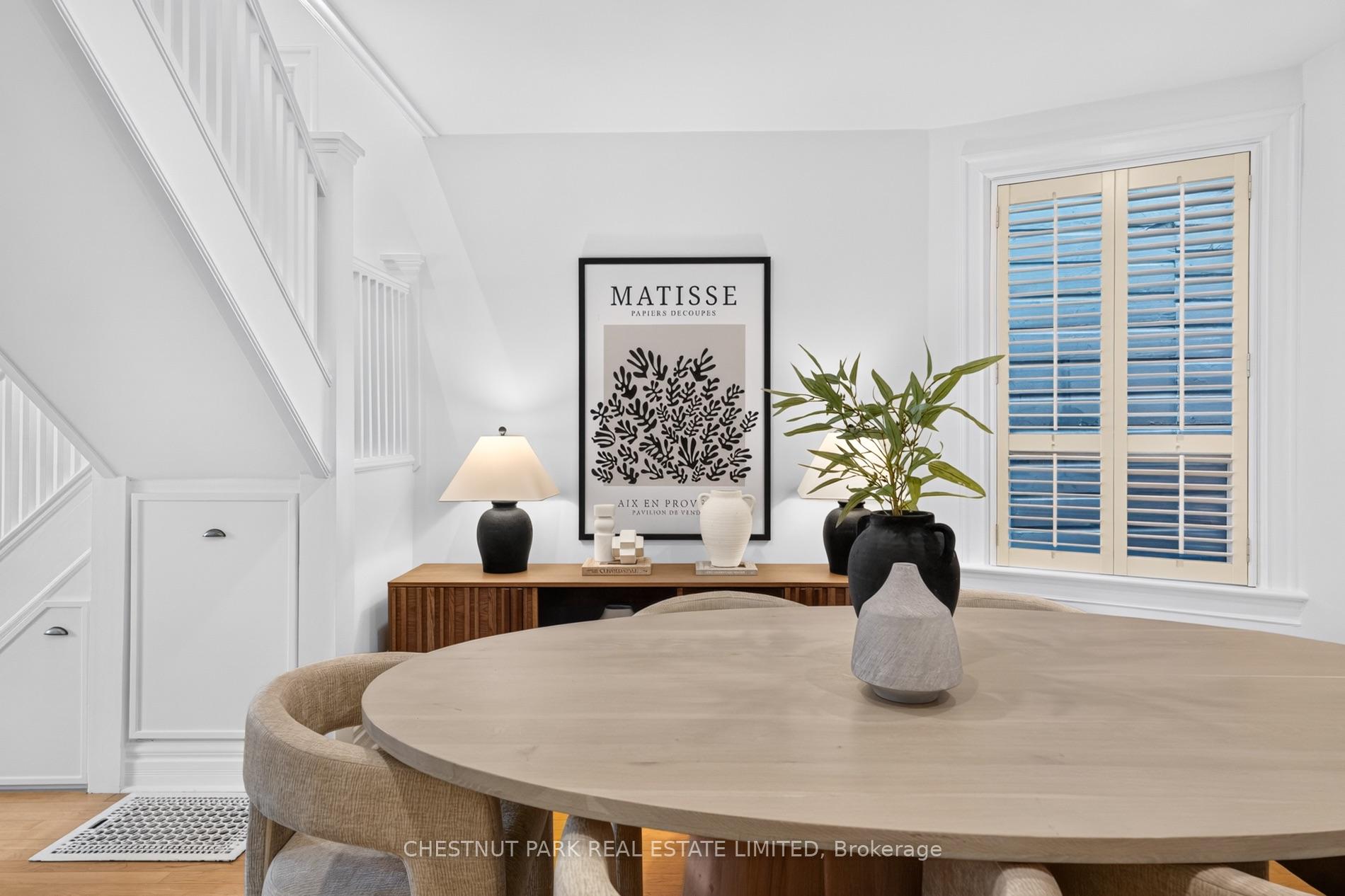 Dining area with a round wood table, white staircase, console, plants, and shuttered window.