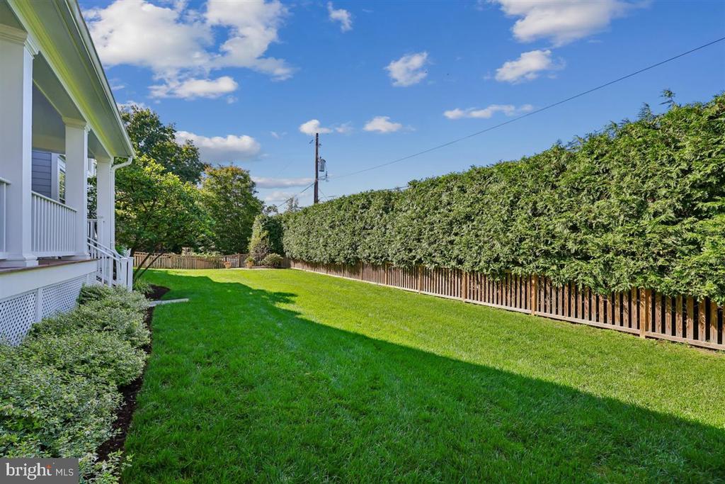 Backyard lawn beside a white porch, bordered by a tall hedge and wooden fence.