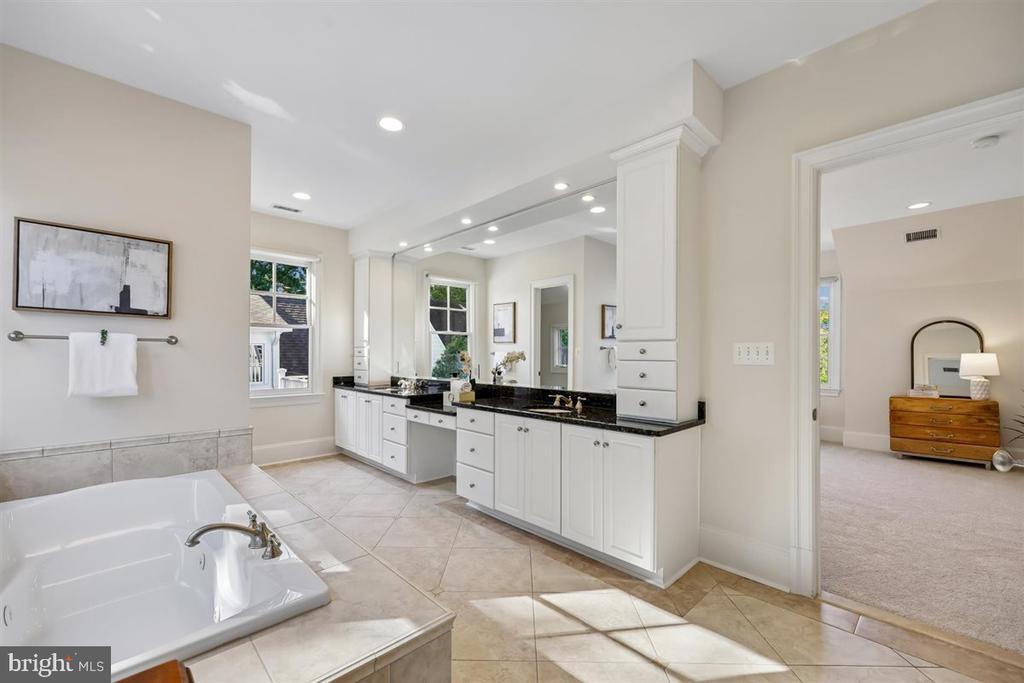 Bathroom with a large soaking tub and long white vanity; furnished bedroom visible through the doorway.