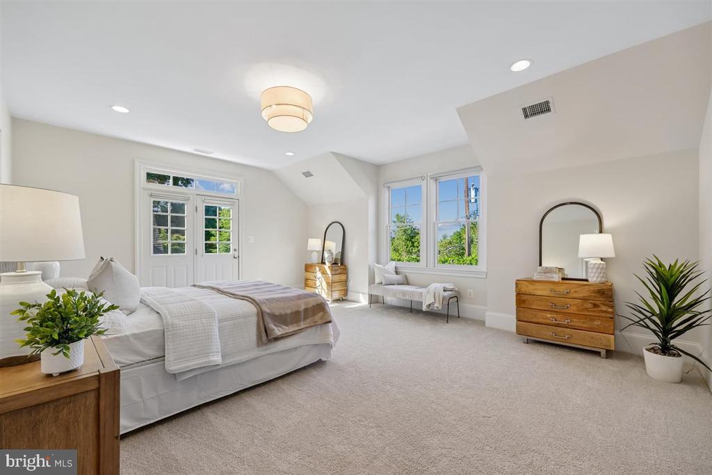 Bright staged bedroom with a large bed, wood dressers, and a bench beneath windows.