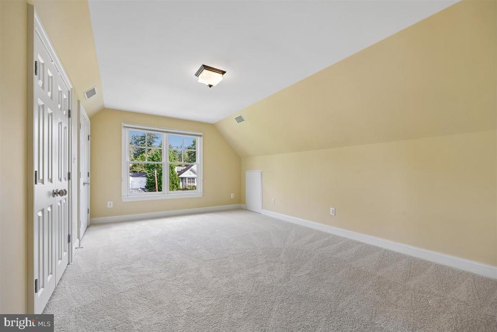 Empty carpeted bedroom with a sloped ceiling and a large window overlooking trees and houses.