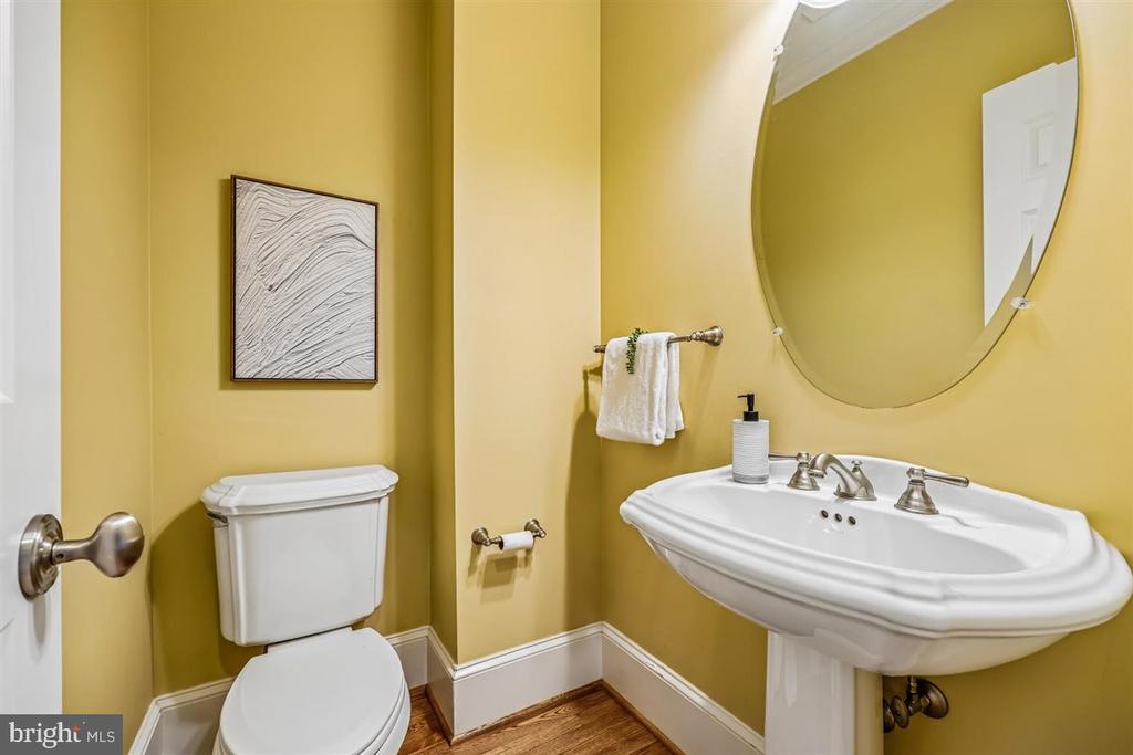 Powder room with mustard-yellow walls, a white pedestal sink, oval mirror, and framed artwork.