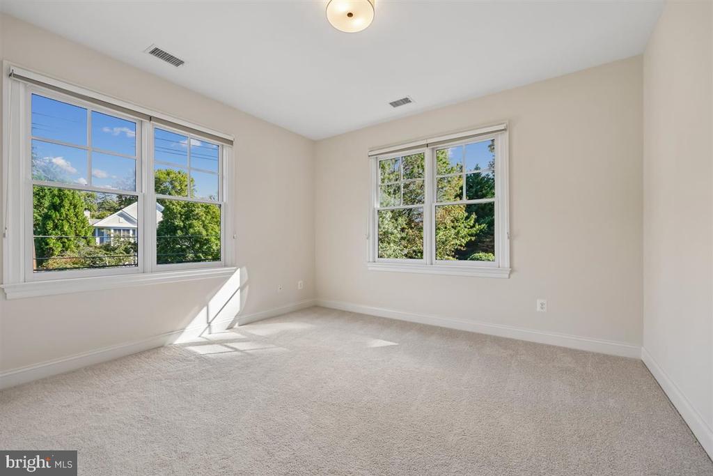 Empty bedroom with beige walls, carpet, and two large windows overlooking trees.