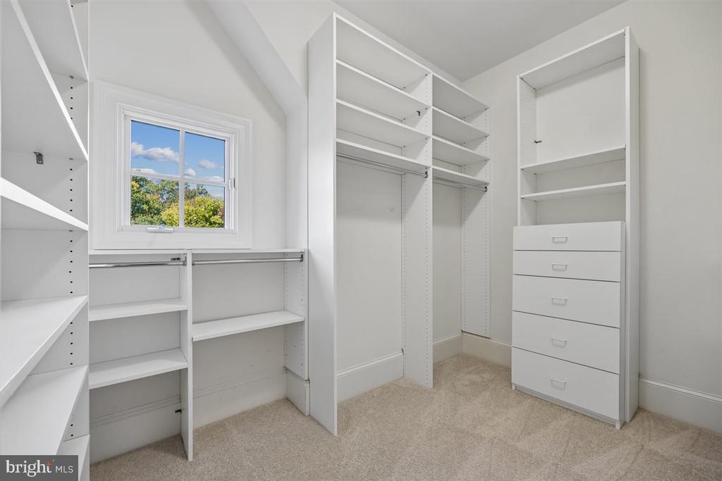 Walk-in closet with white built-in shelves, hanging rods, drawers, and a window overlooking trees.