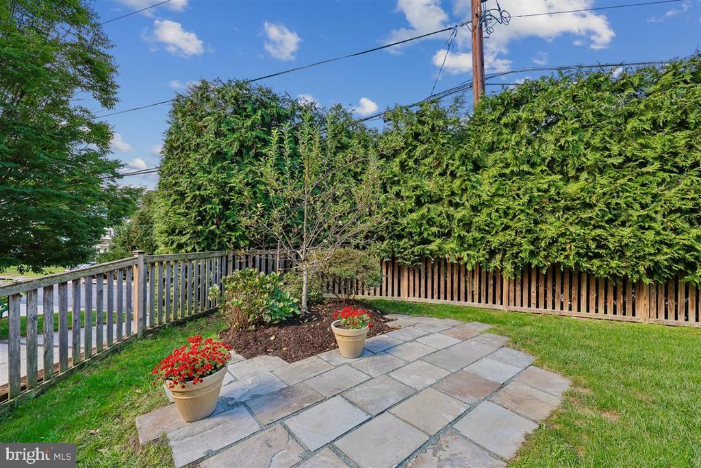 Stone patio bordered by tall hedges and wooden fences, with potted flowers.