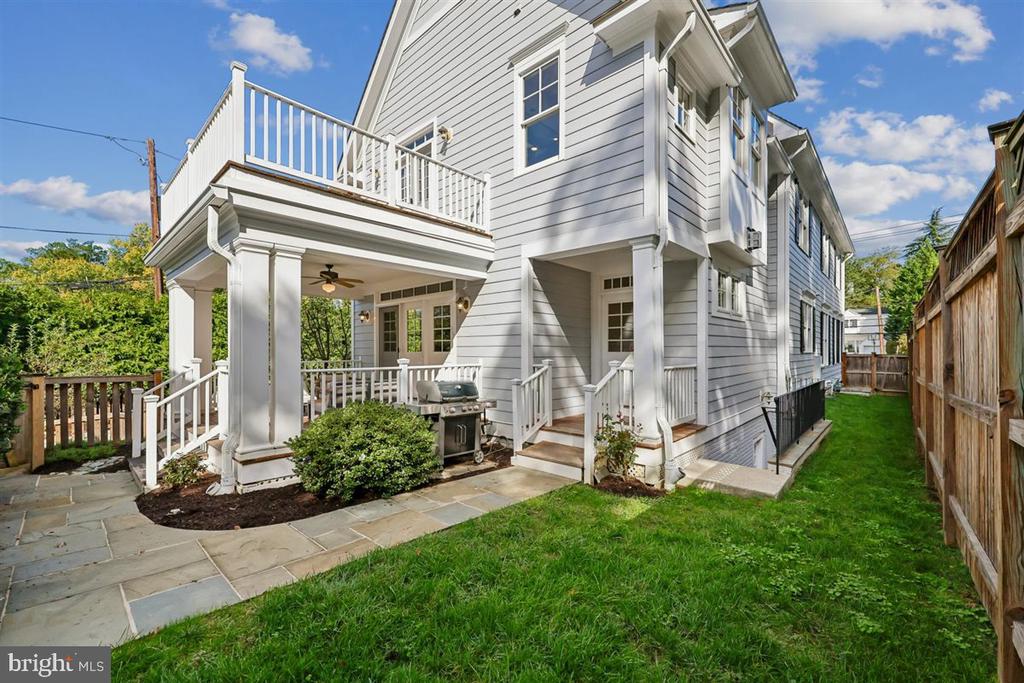 Rear of a gray two-story home with a balcony, covered patio, grill, stone paving, and fenced lawn.