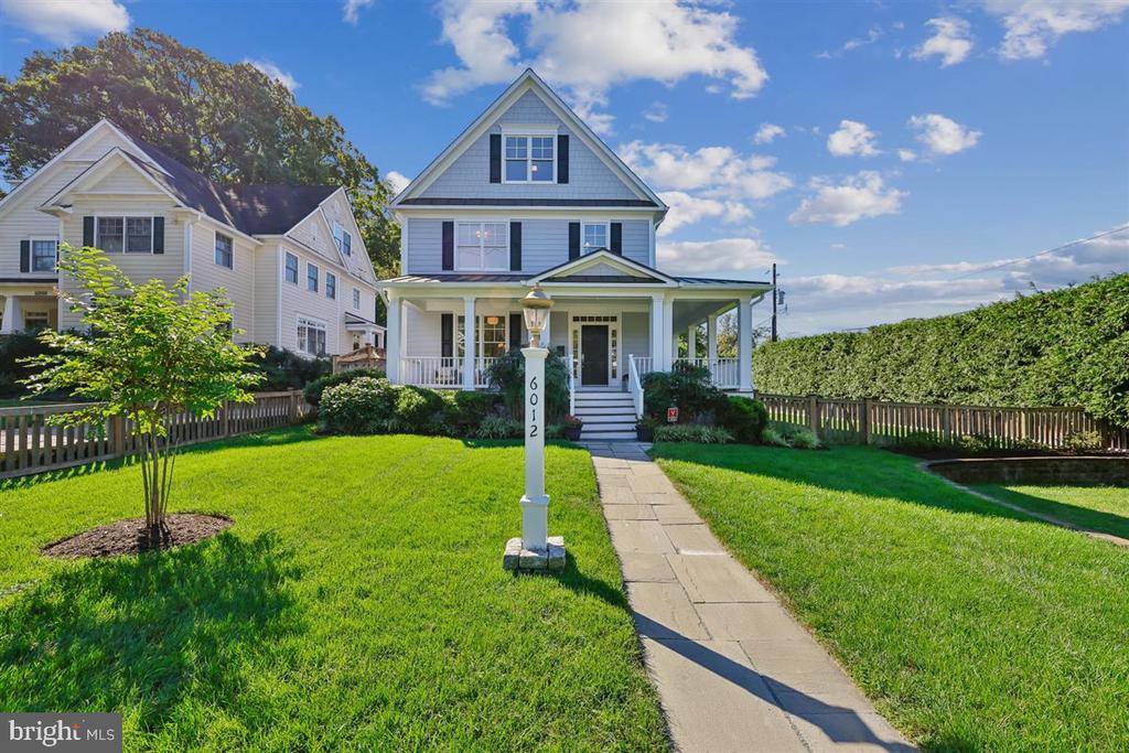 Two-story blue-gray house with a covered porch and walkway across a sunny front lawn.