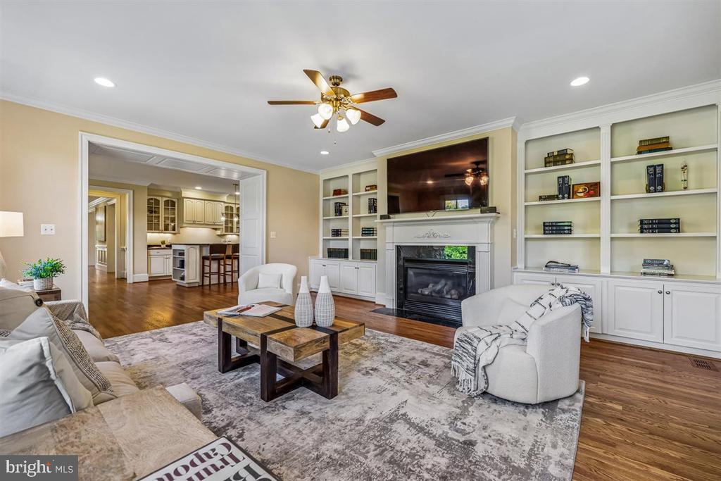 Living room with built-in bookcases, fireplace, television, white seating, and an open view into the kitchen.
