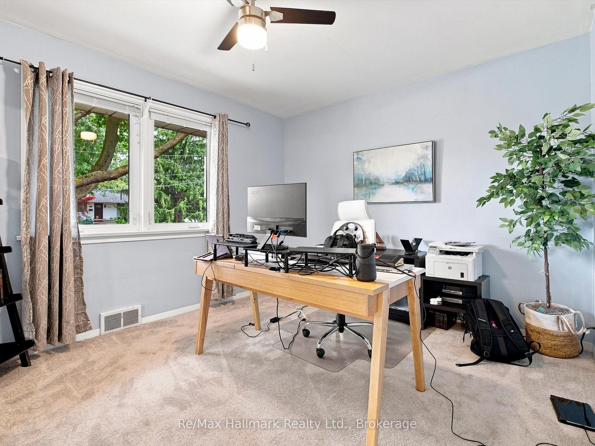 Home office with a wooden desk, computer equipment, printer, and a large window overlooking trees.
