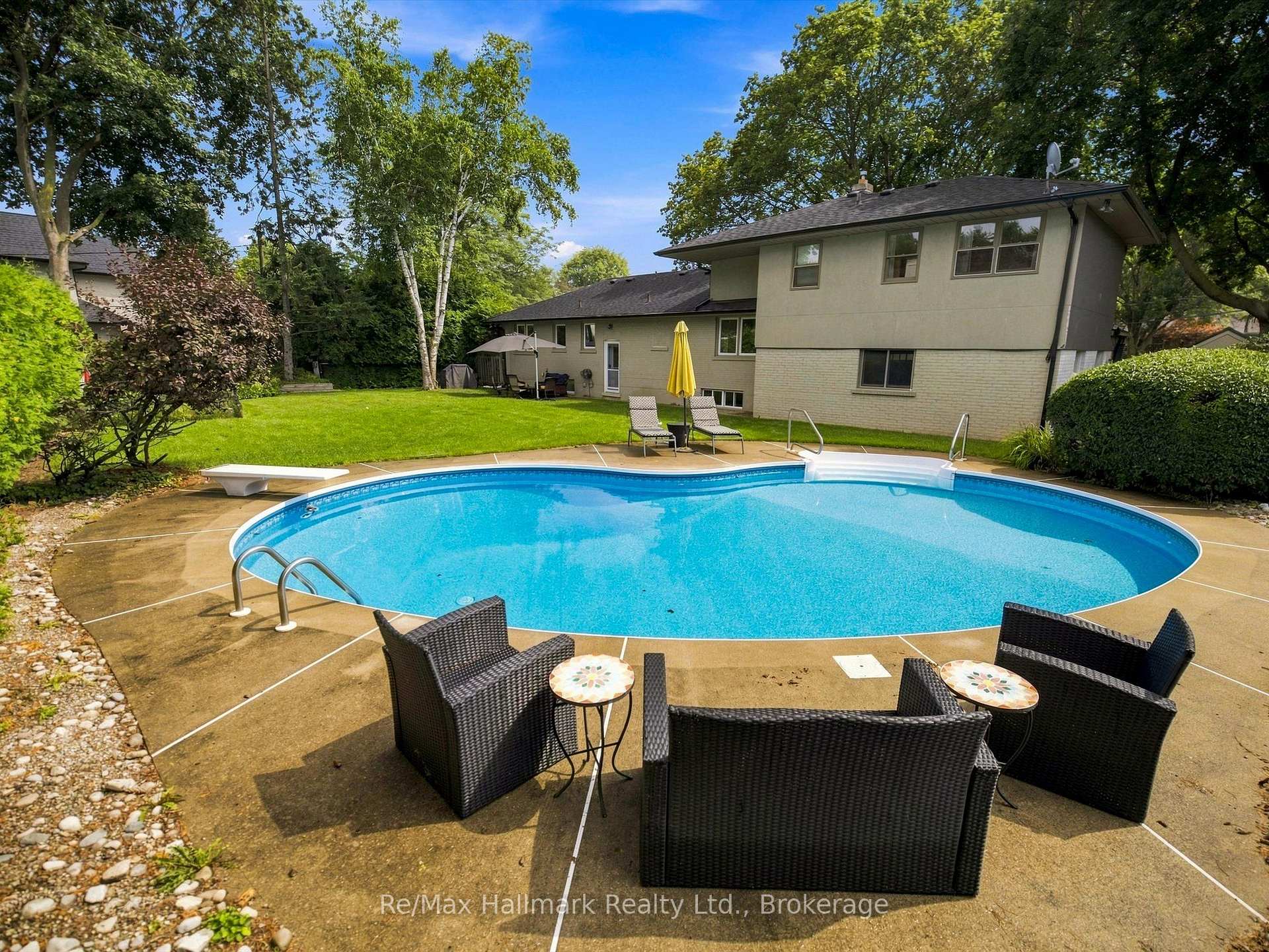 Backyard swimming pool beside a two-storey house, with patio seating and mature trees.